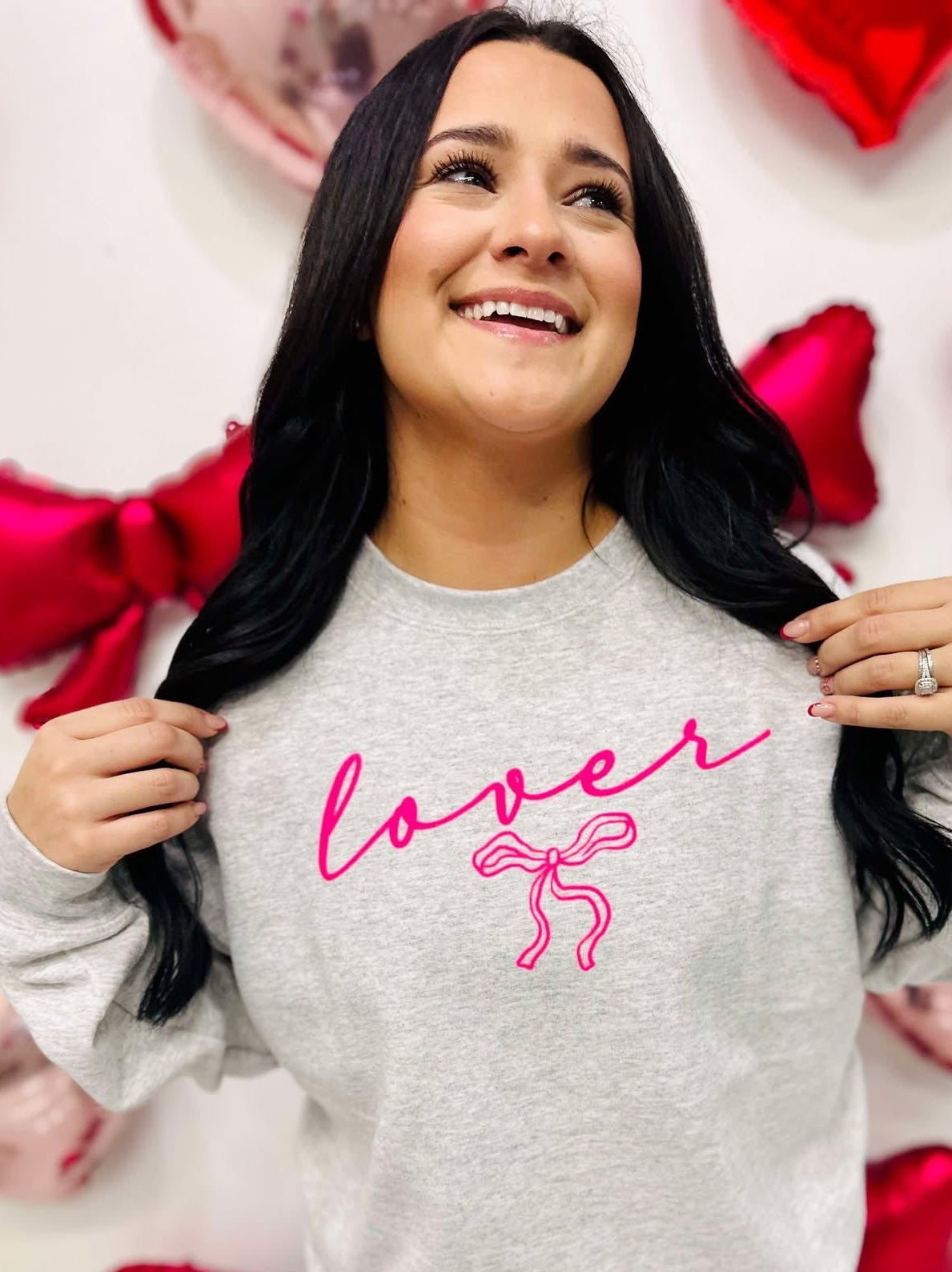 A woman smiling and posing with a gray sweatshirt that has the word 'lover' written in neon pink and a pink bow graphic.
