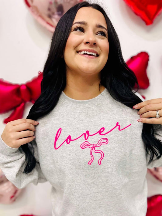 A woman smiling and posing with a gray sweatshirt that has the word 'lover' written in neon pink and a pink bow graphic.