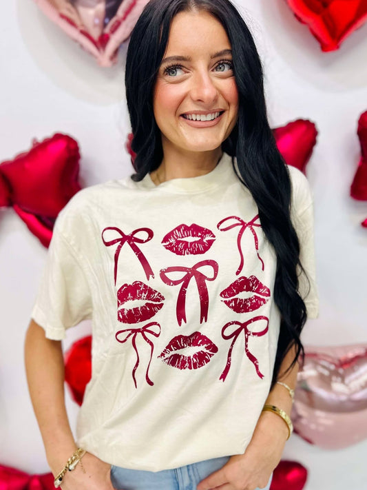 A person wearing a beige t-shirt with a graphic print of three stylized lips and bows in red and white colors, standing against a backdrop of Valentine's Day-themed decorations including balloons and hearts.