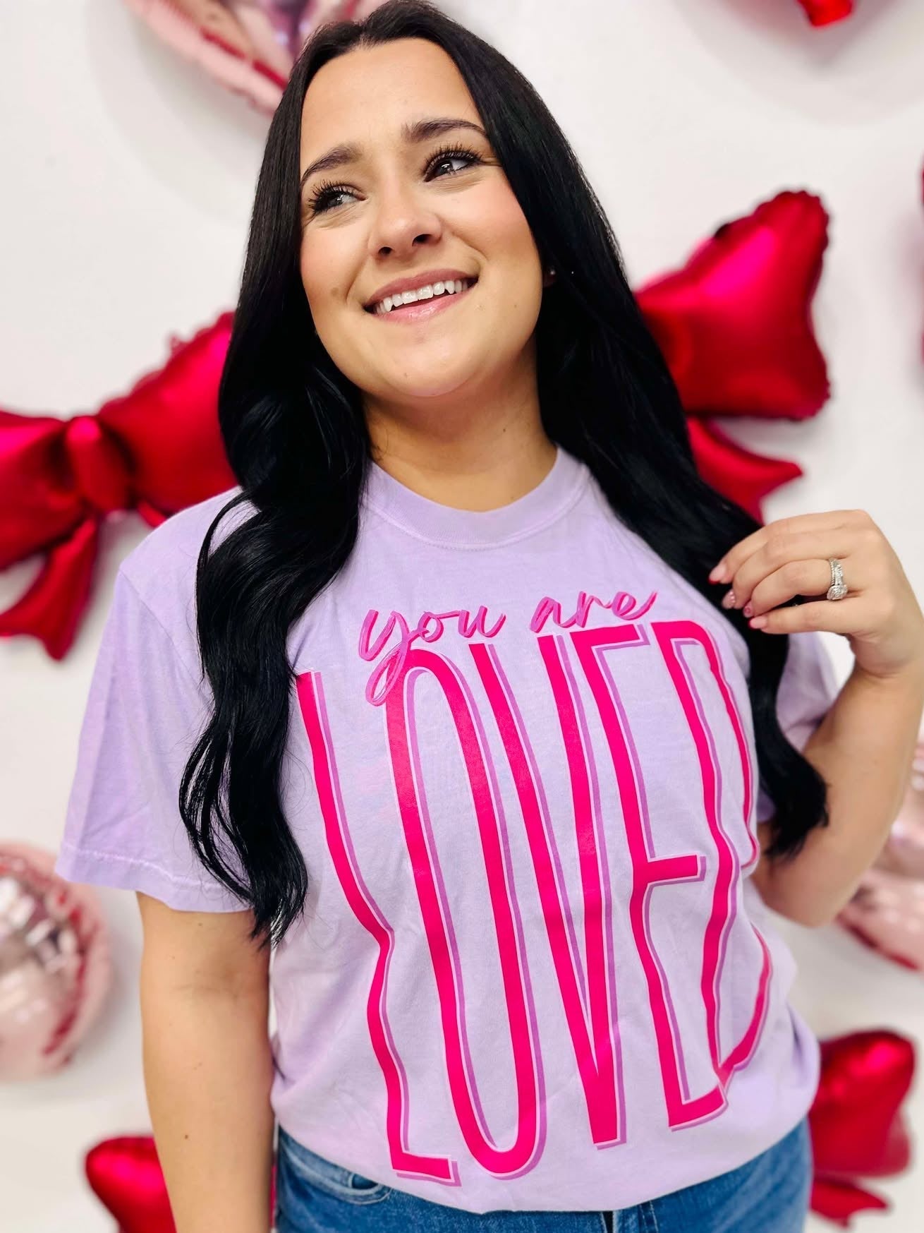 A woman smiling and wearing a purple t-shirt with the text 'you are LOVED' printed in large pink letters.