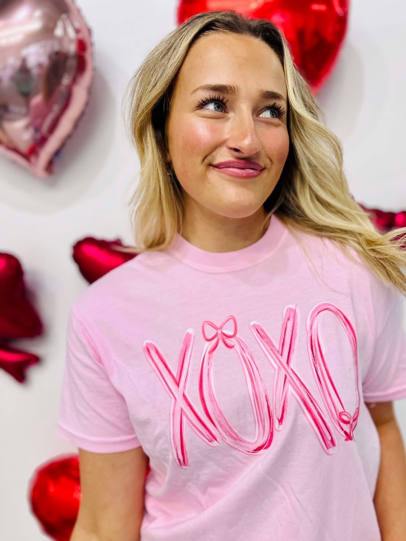 A woman wearing a pink t-shirt with the text 'XOXO' and a pink bow design on it, standing in a room with balloons and decorative hearts.