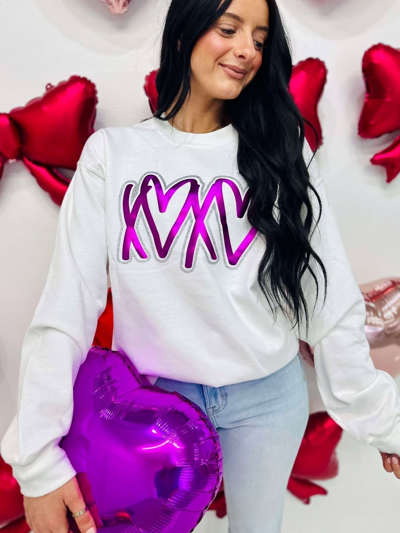 Woman wearing a white sweatshirt with purple text, holding a purple heart-shaped balloon, standing against a wall with red heart decorations.