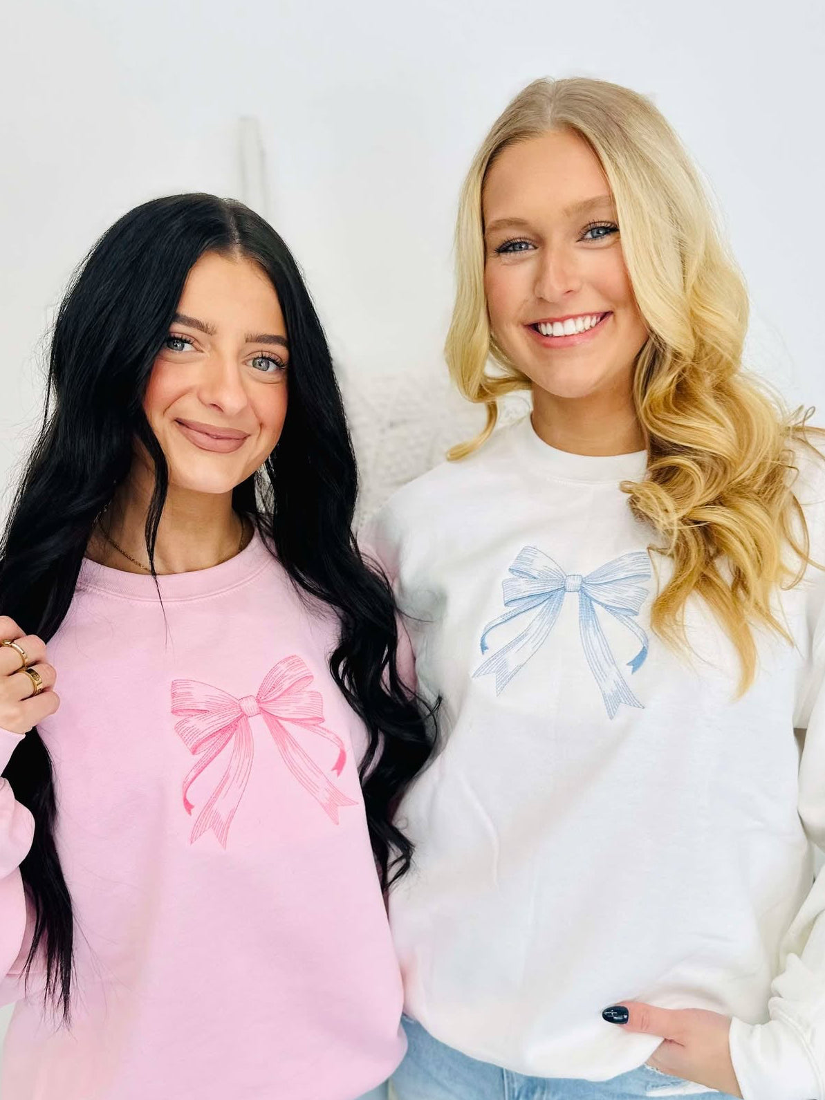 Two individuals standing next to each other, one wearing a pink and the other wearing a white embroidered bow sweatshirt.