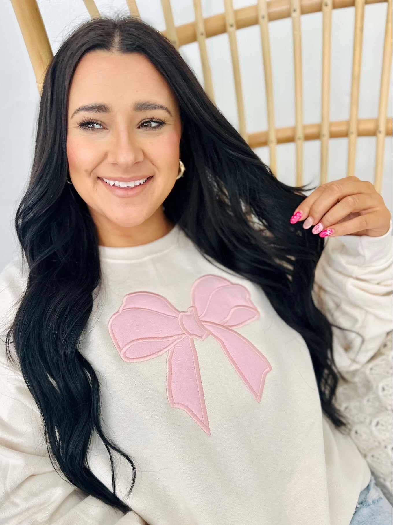 A woman smiling and posing in a beige sweatshirt with a pink bow applique on the front.