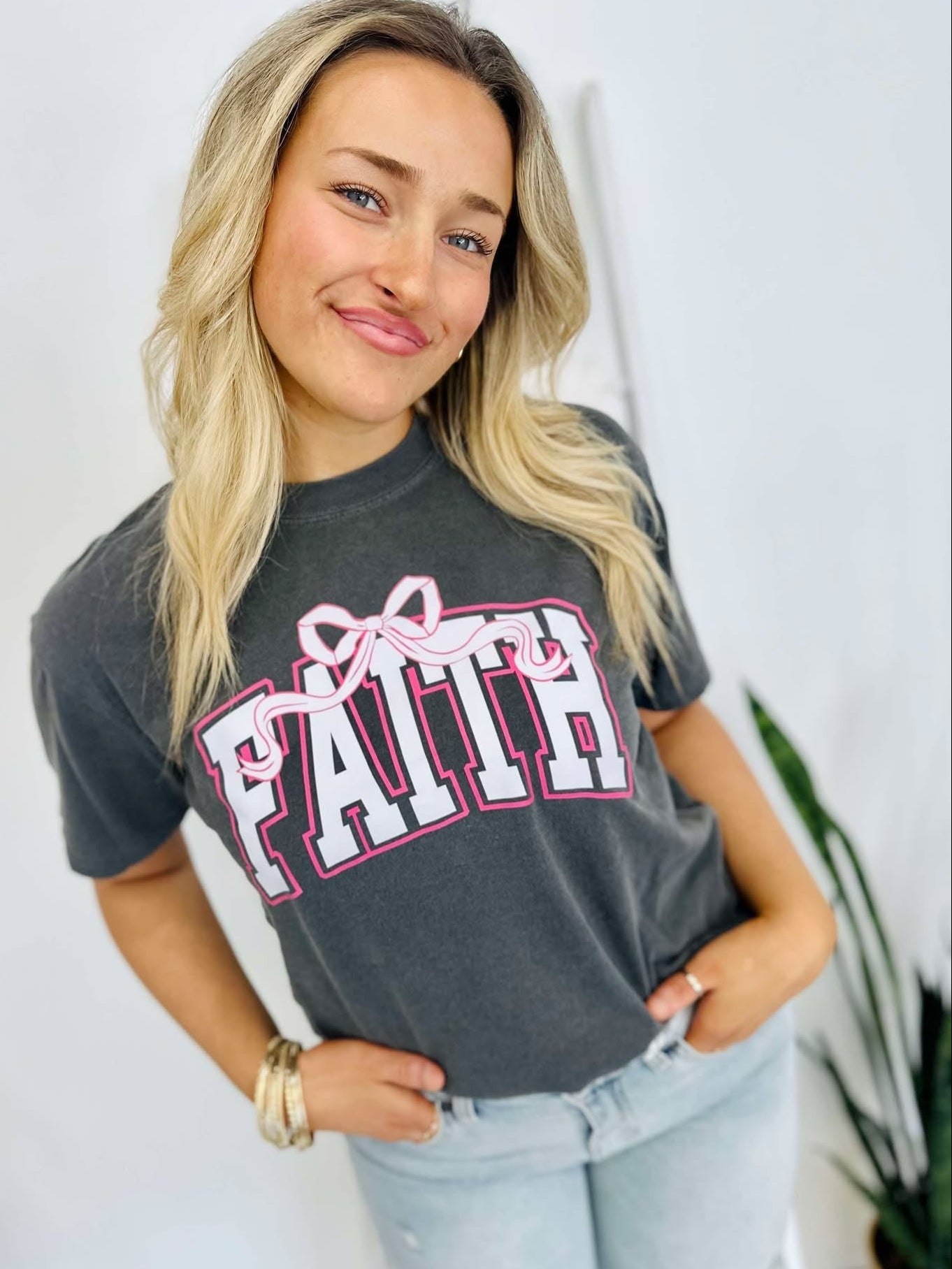 A woman wearing a gray t-shirt with the word 'Faith' and a pink bow on it, standing with one hand against a white background.