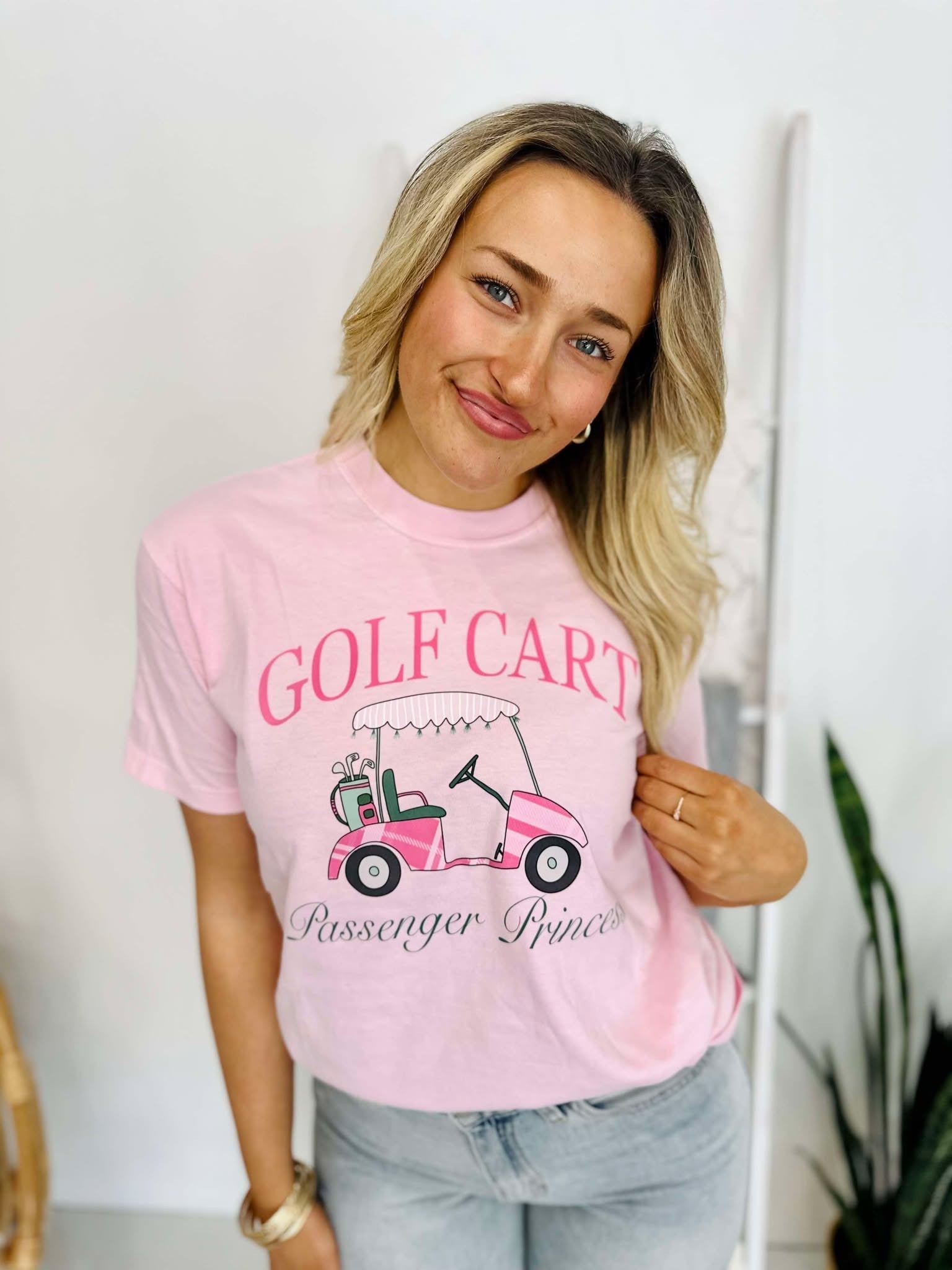 A woman wearing a pink t-shirt with 'GOLF CART' and 'PASSENGER PRINCESS' printed on it, standing in a room with a white background.