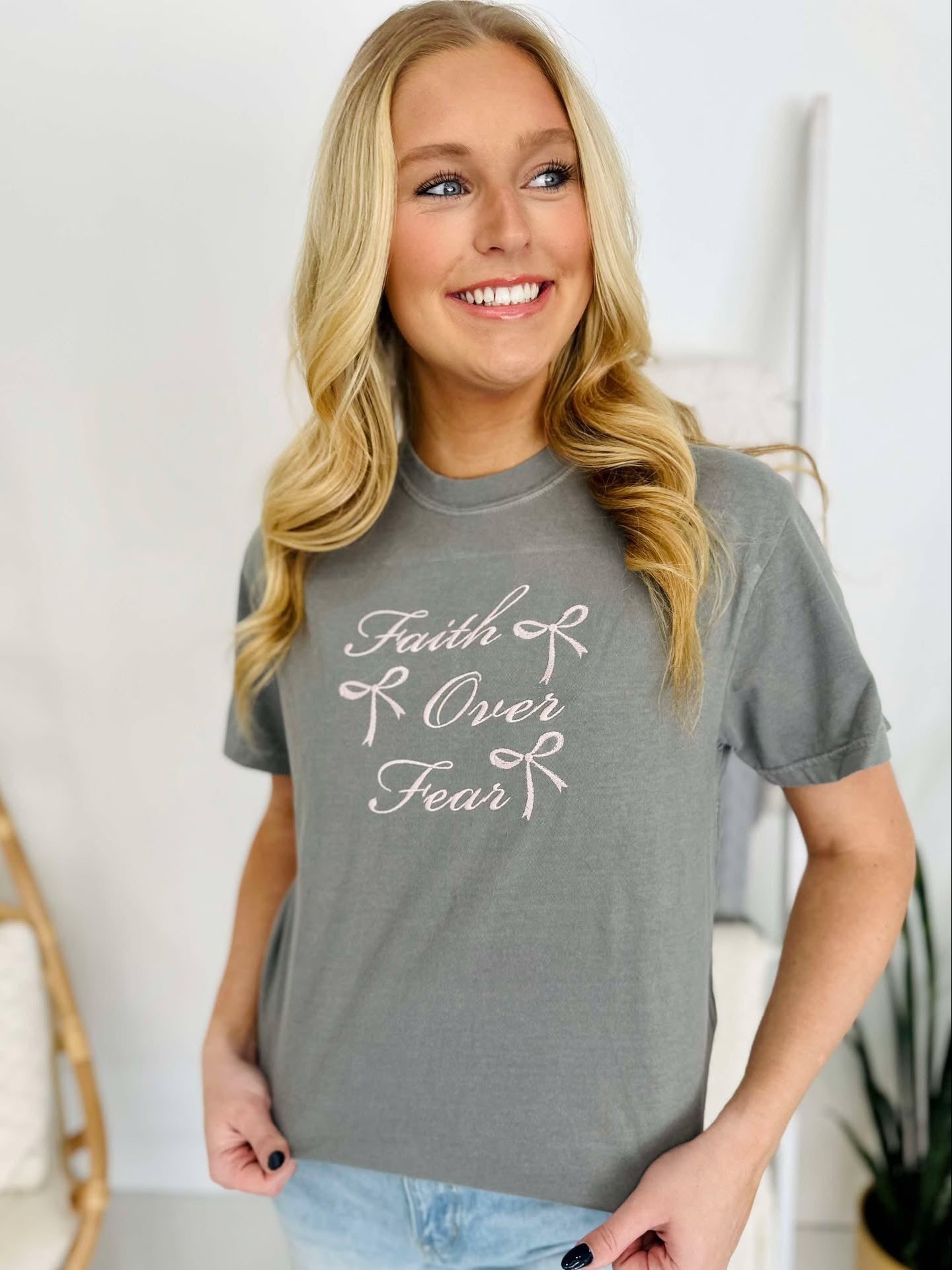 A woman smiling and wearing a gray t-shirt with 'Faith & Fear' embroidered on the front.
