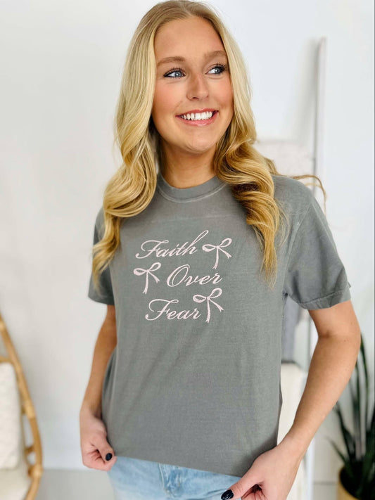 A woman smiling and wearing a gray t-shirt with 'Faith & Fear' embroidered on the front.