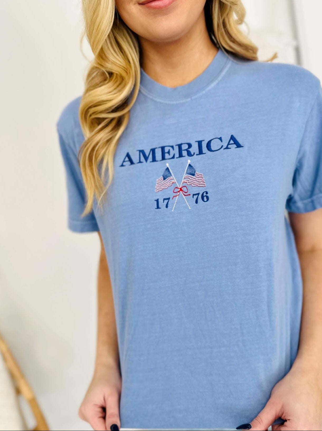 A woman wearing a blue t-shirt with 'America 1776' and a flag-themed design embroidered on the front.