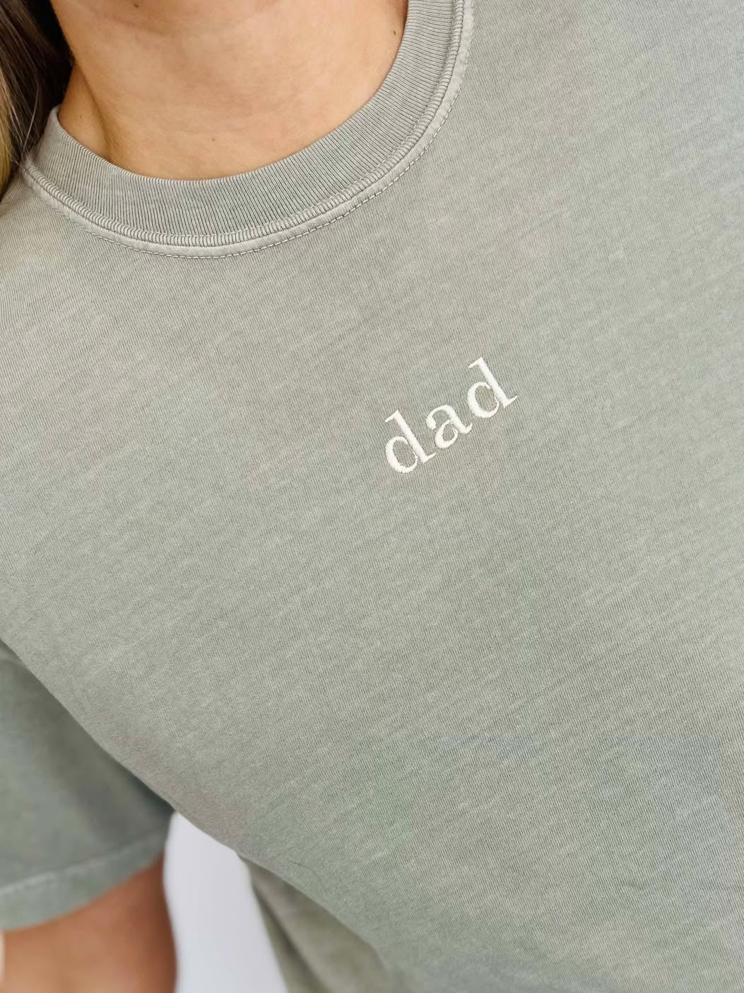 A person wearing a sandstone-colored t-shirt with the word 'DAD' embroidered on the front.