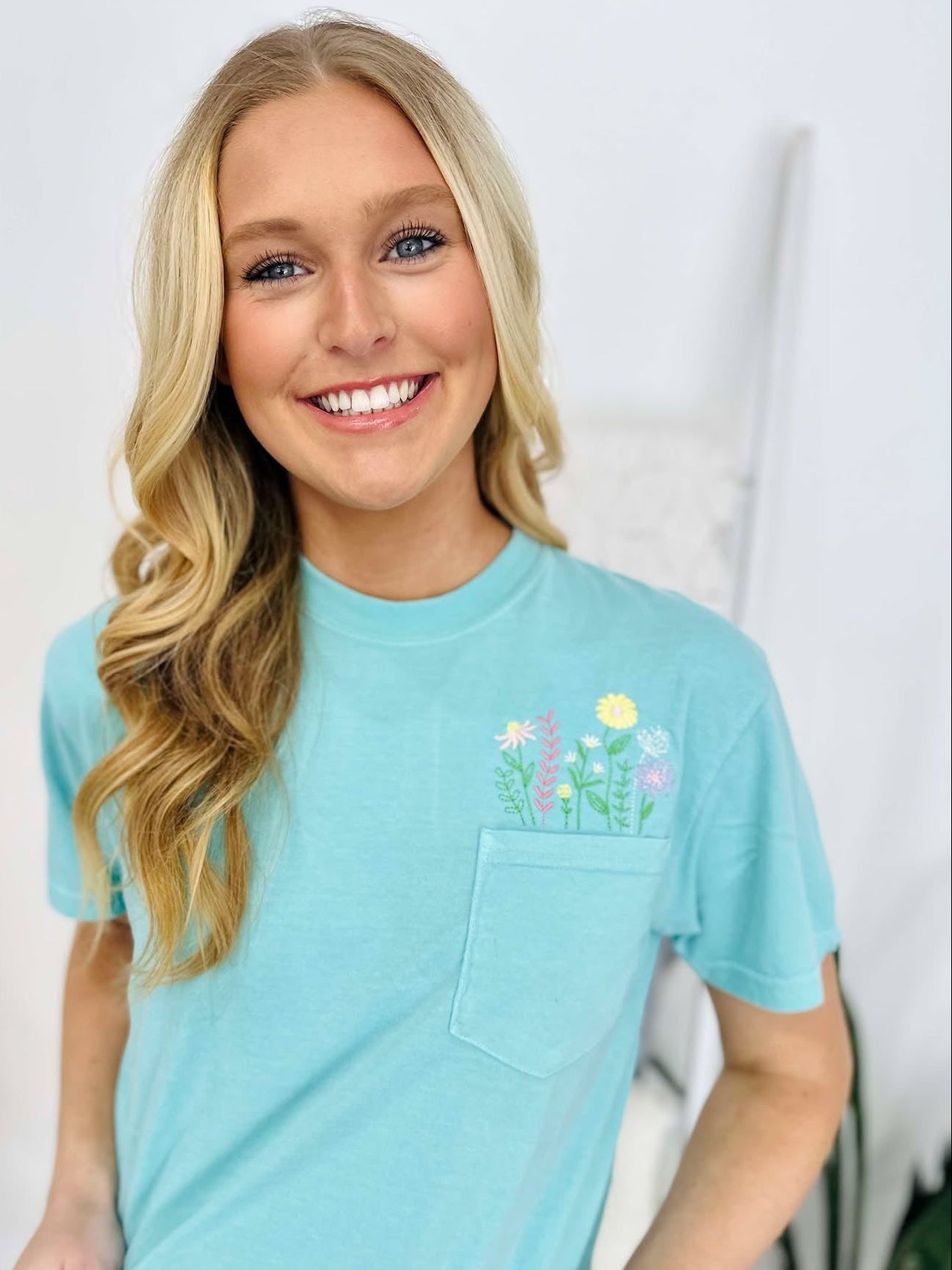 A woman wearing a chalky mint green t-shirt with a floral pocket design and embroidered lettering on the front, smiling against a white background.
