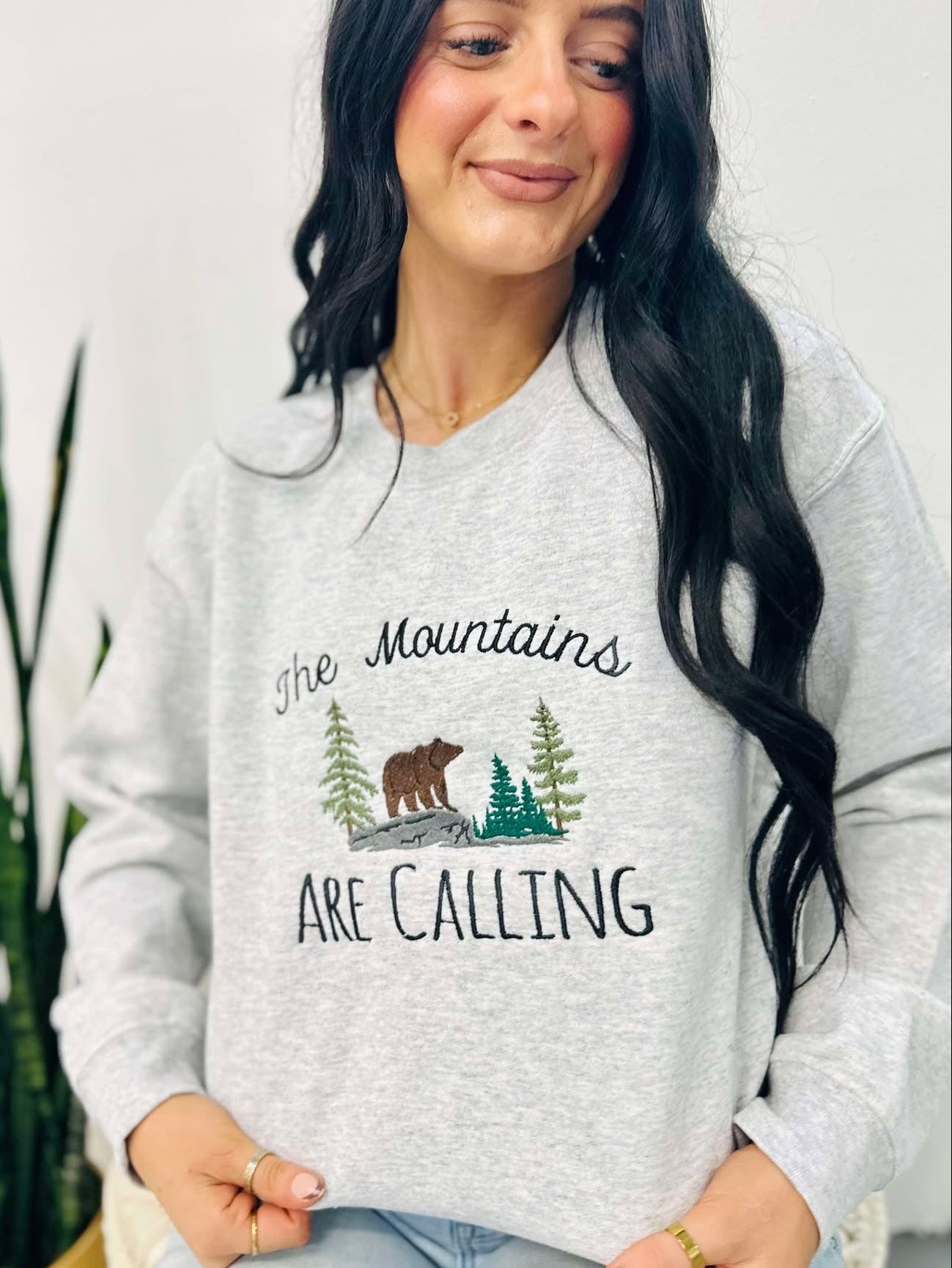 A woman smiling, wearing an ash grey embroidered sweatshirt with the text 'THE MOUNTAINS ARE CALLING' and an image of a bear in a forest.