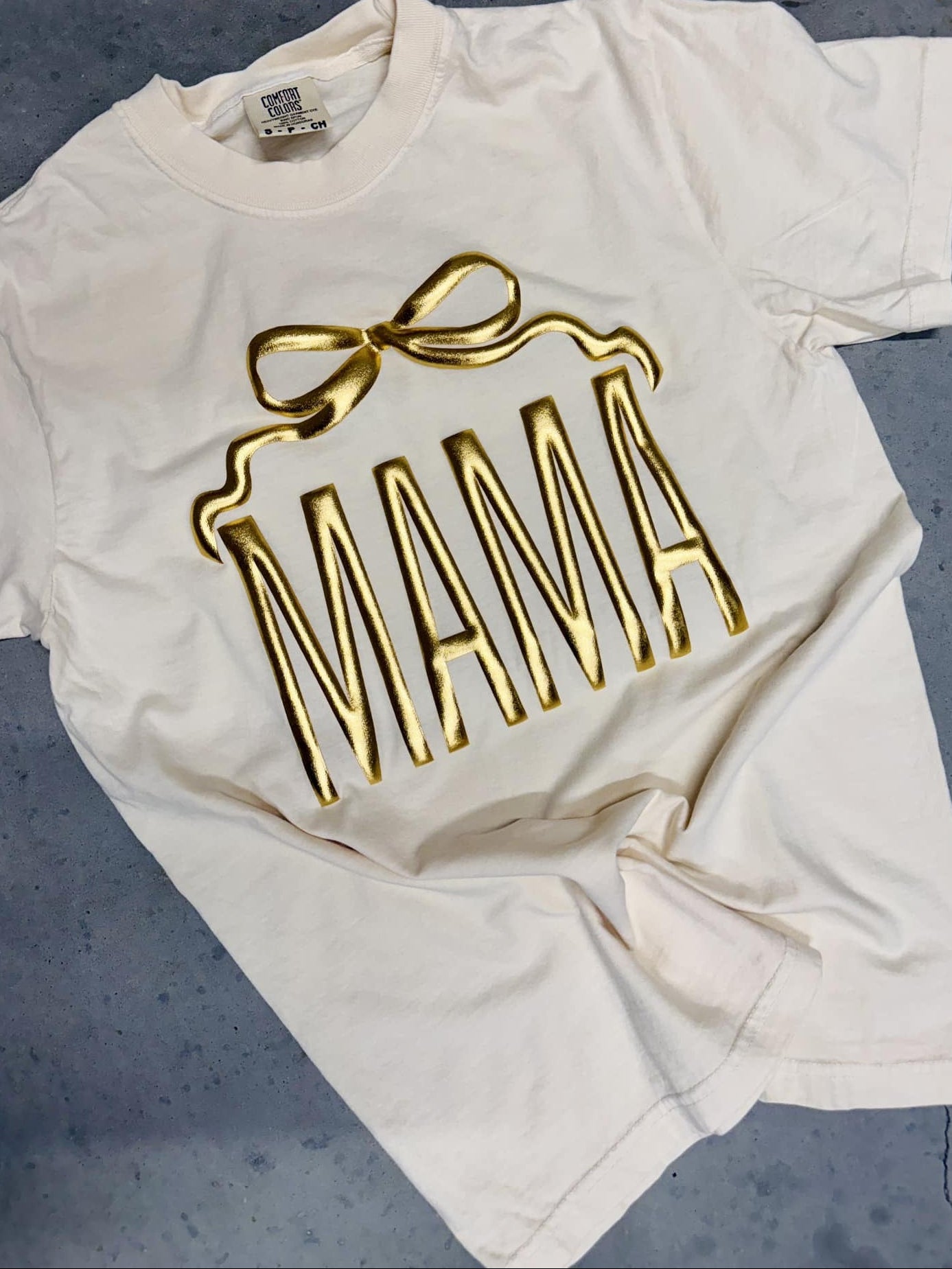 An ivory colored t-shirt with the word 'MAMA' printed in gold metallic vinyl in a bow shape.