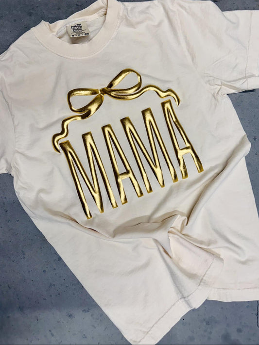 An ivory colored t-shirt with the word 'MAMA' printed in gold metallic vinyl in a bow shape.
