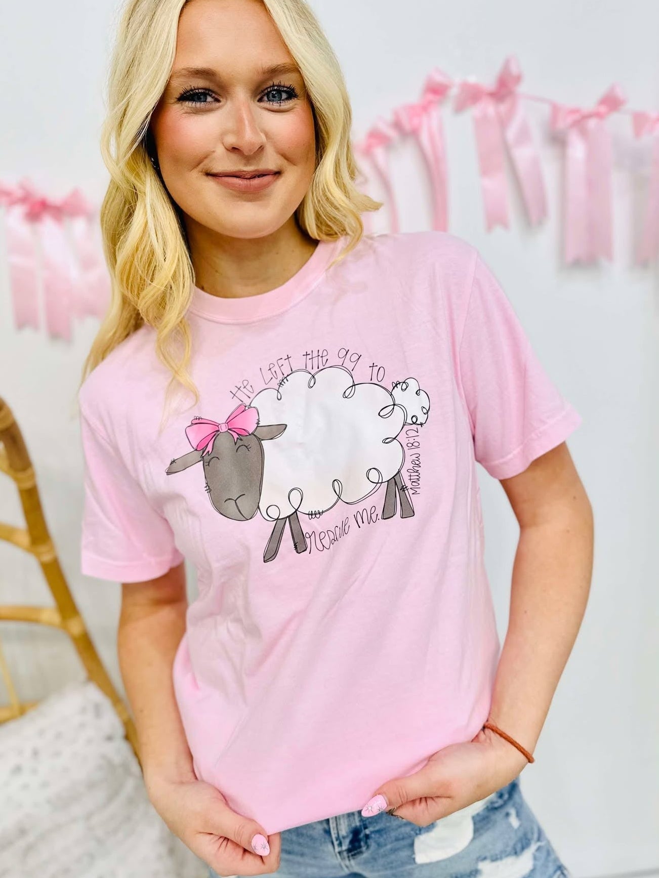 A person wearing a pink graphic tee with text and a sheep design, standing in front of a white background with decorative elements.