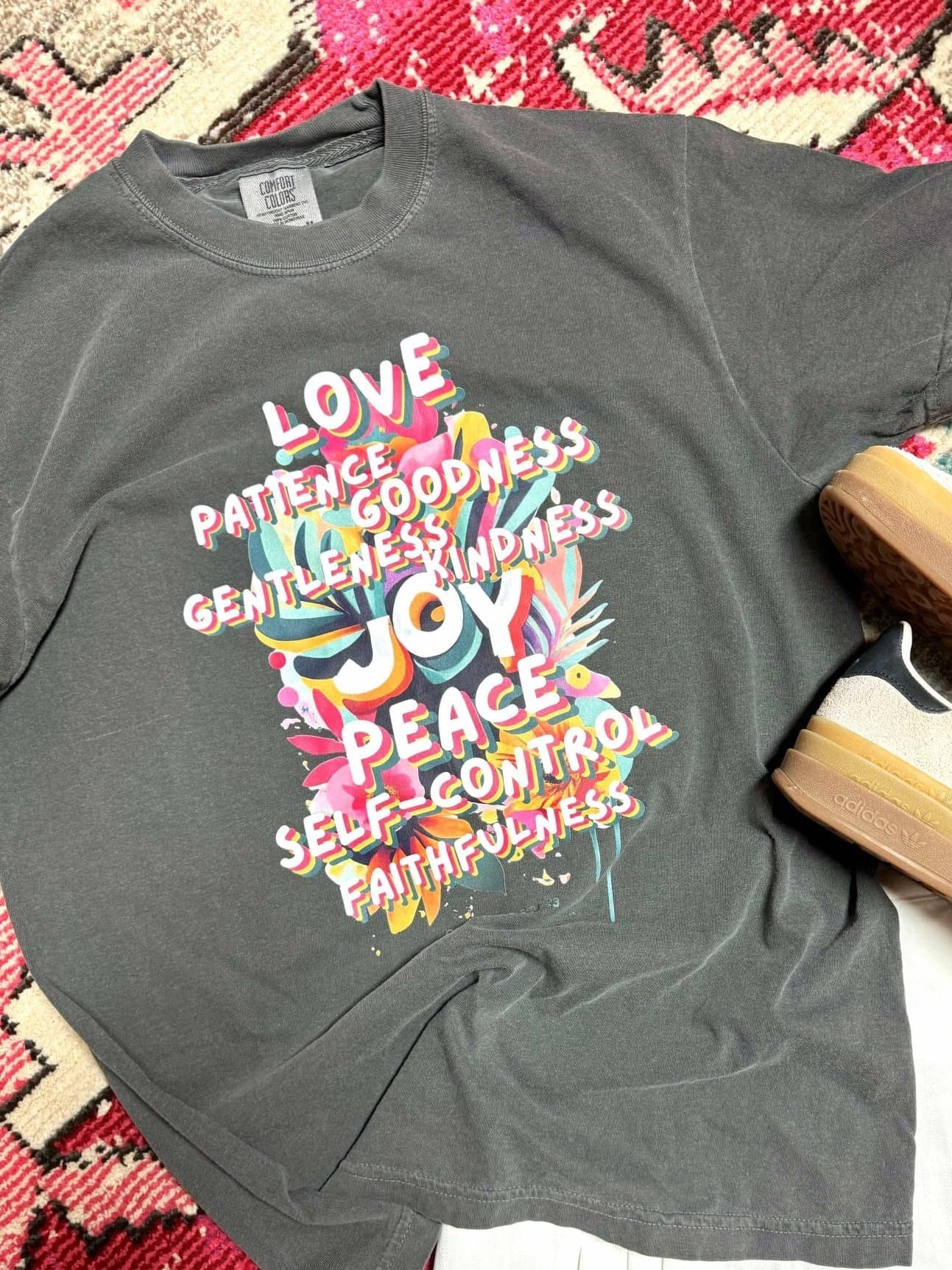 A gray t-shirt laid out on a patterned surface, featuring a printed design with the fruits of the spirit, accompanied by a pair of shoes.