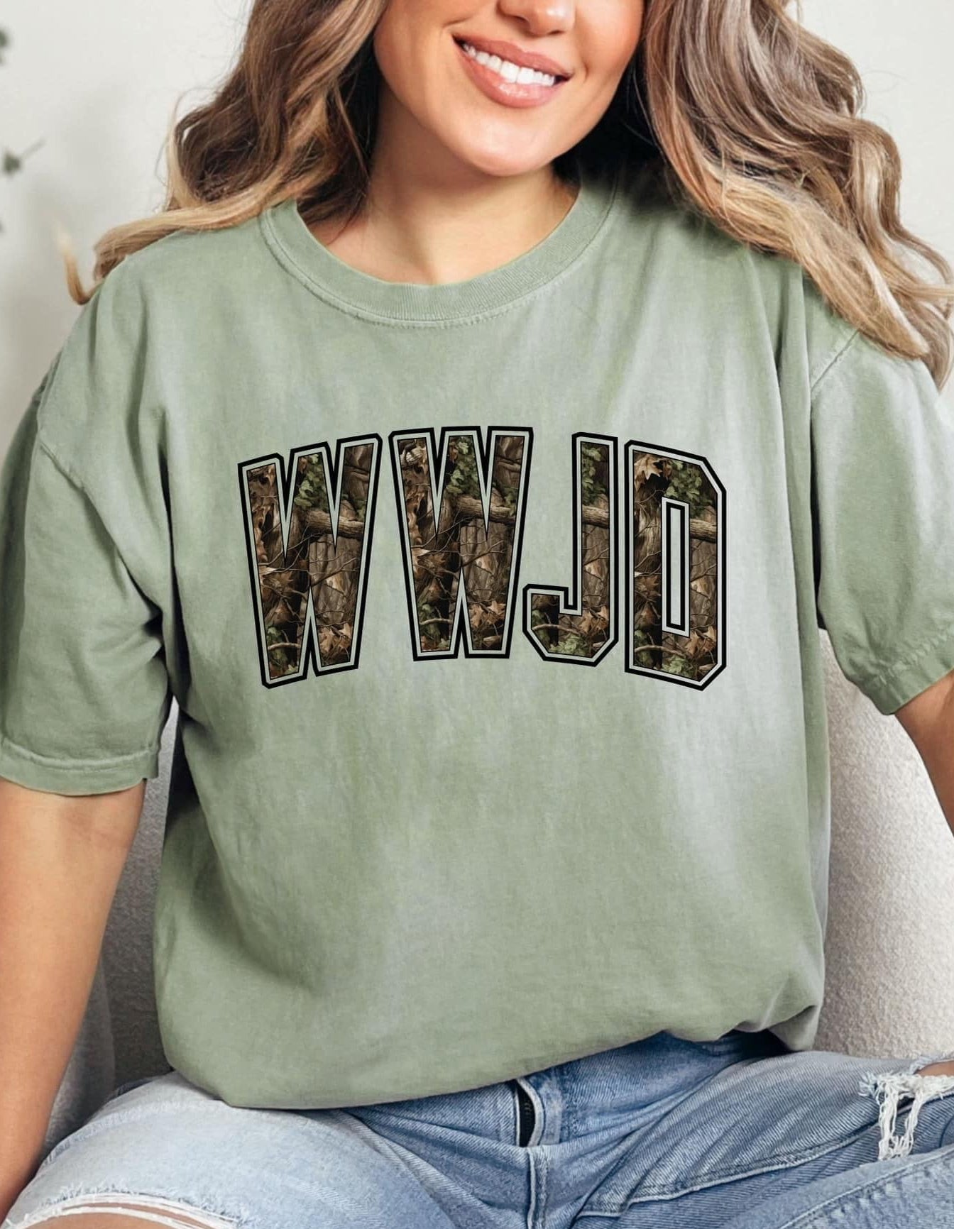 A person wearing a green t-shirt with 'WWJD' camo graphic text on it, sitting on a surface with blue jeans.