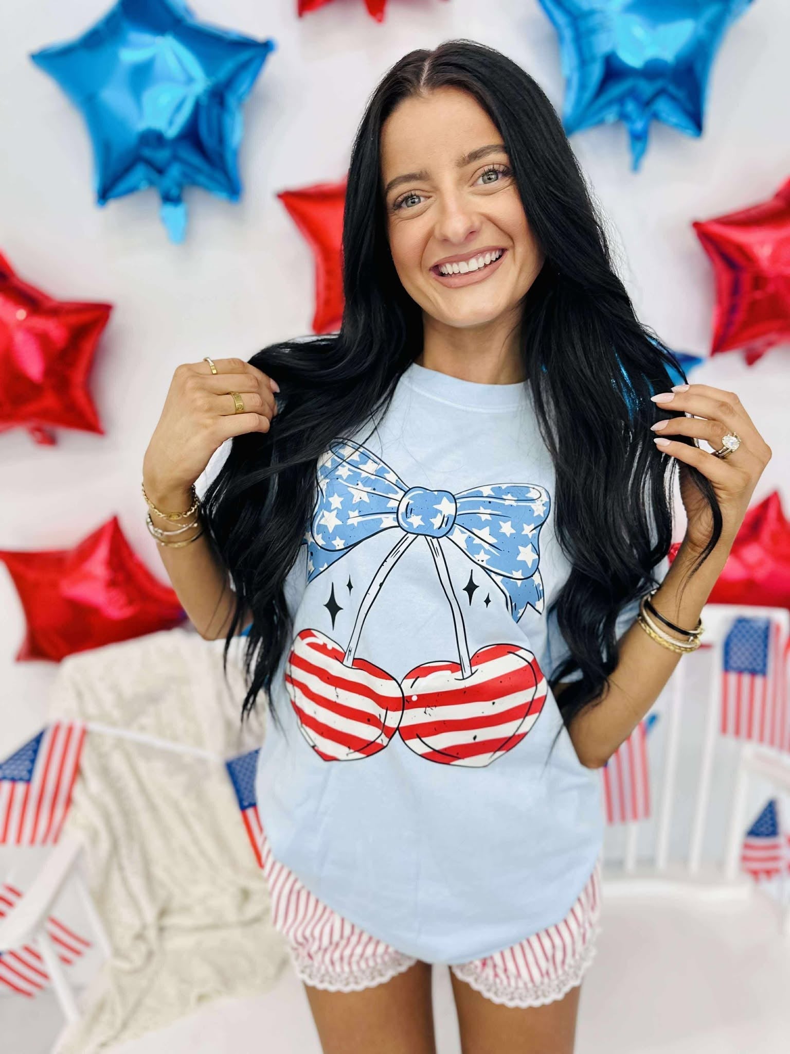 A person wearing a light blue graphic tee with a patriotic cherry design, shorts, and standing in front of a backdrop with red and blue stars.