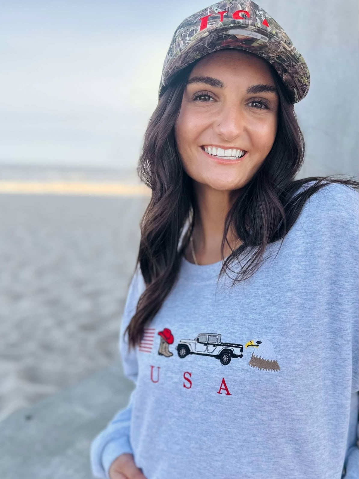 Woman wearing a light blue sweatshirt with an eagle, cowboy boots, truck and usa flag graphic design, sitting on a beach.