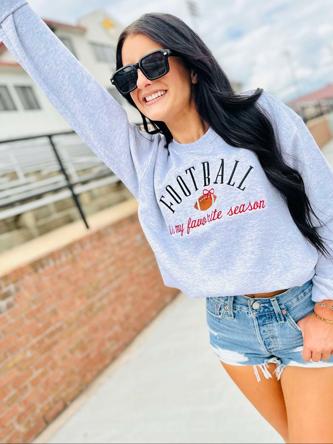 Woman wearing a gray sweatshirt with 'Football is my favorite season' text, sunglasses, and denim shorts outdoors.