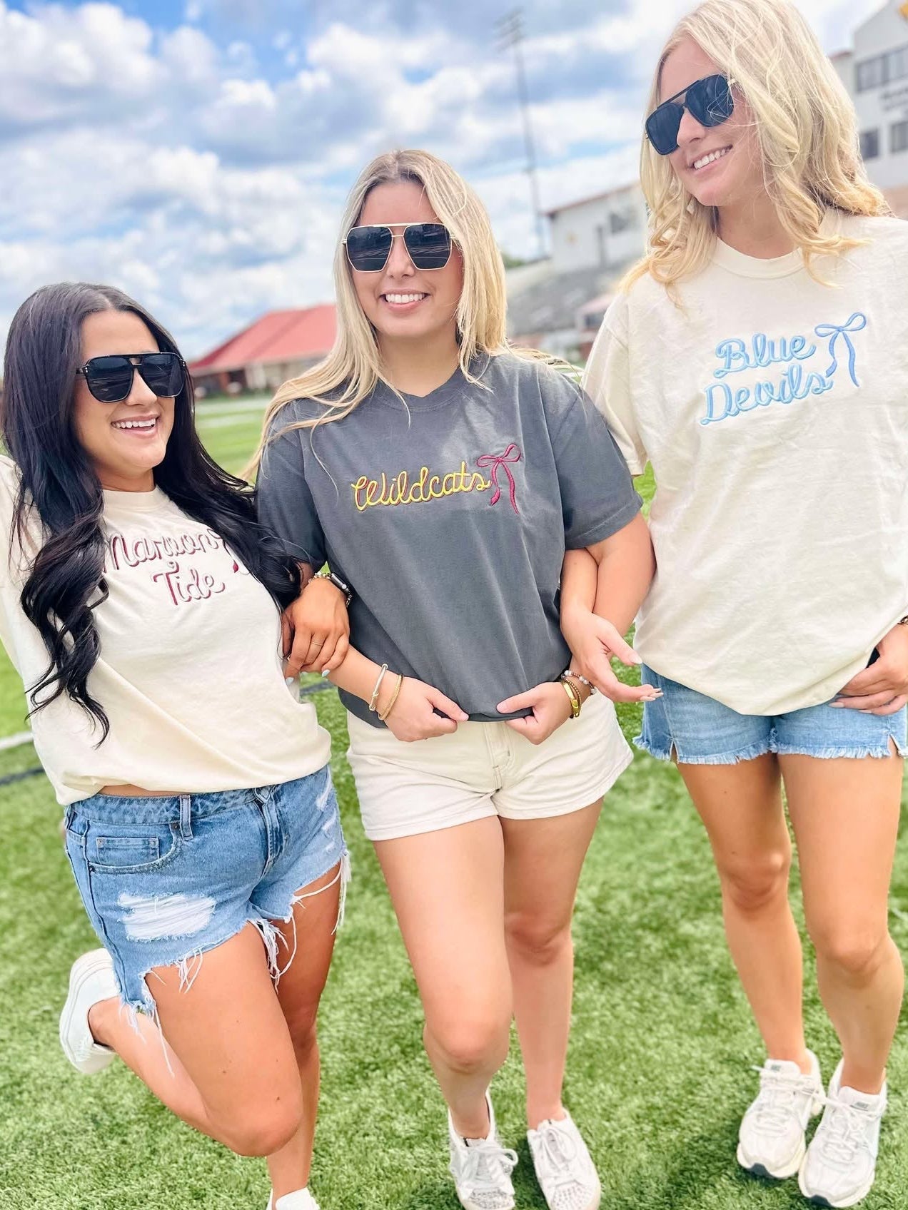 Three women standing on a grassy field wearing casual outfits with visible mascot school  text on their shirts.