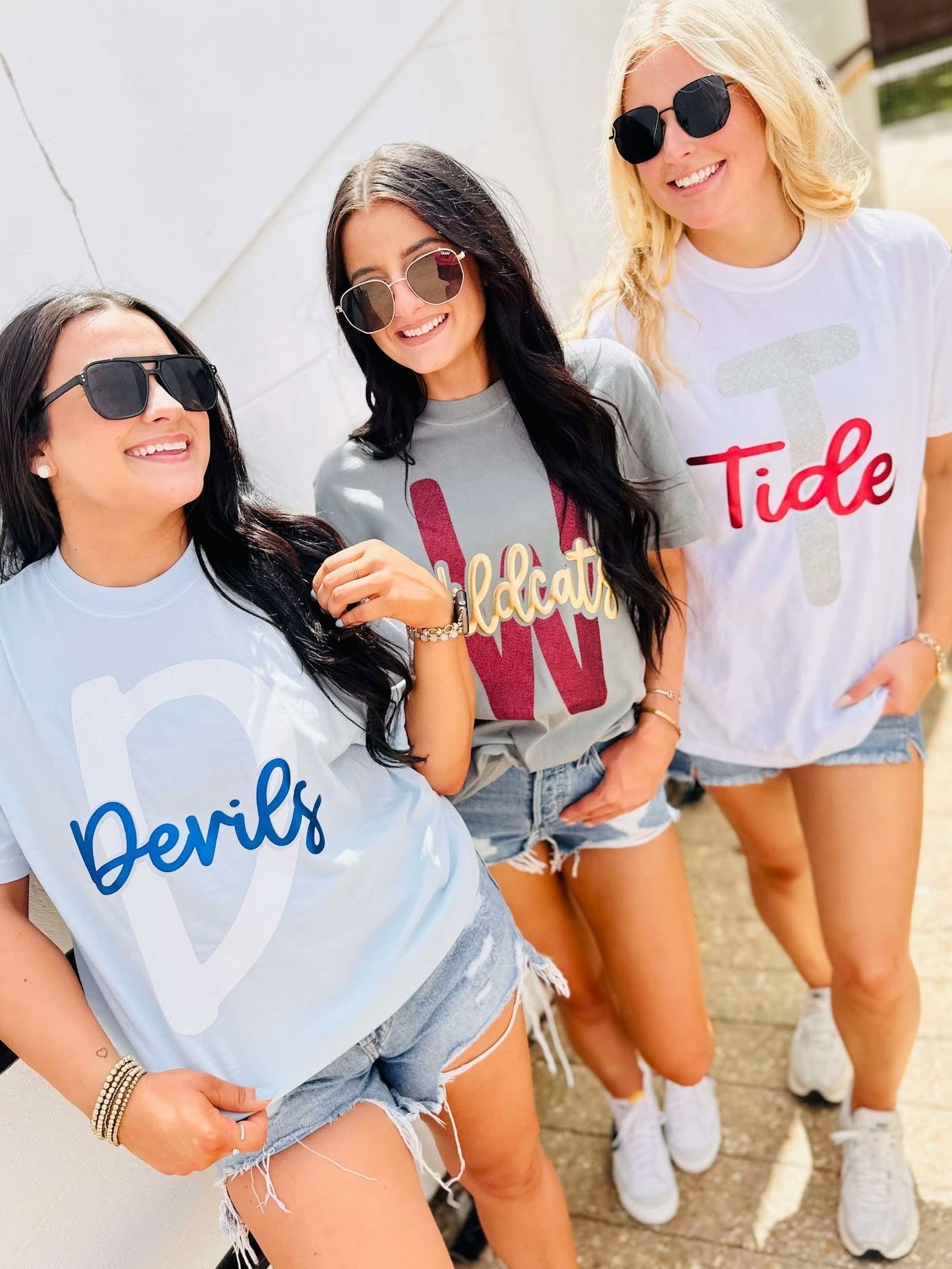 Three women wearing t-shirts with 'Devils', 'W', and 'Tide' glitter puff text, standing outdoors.