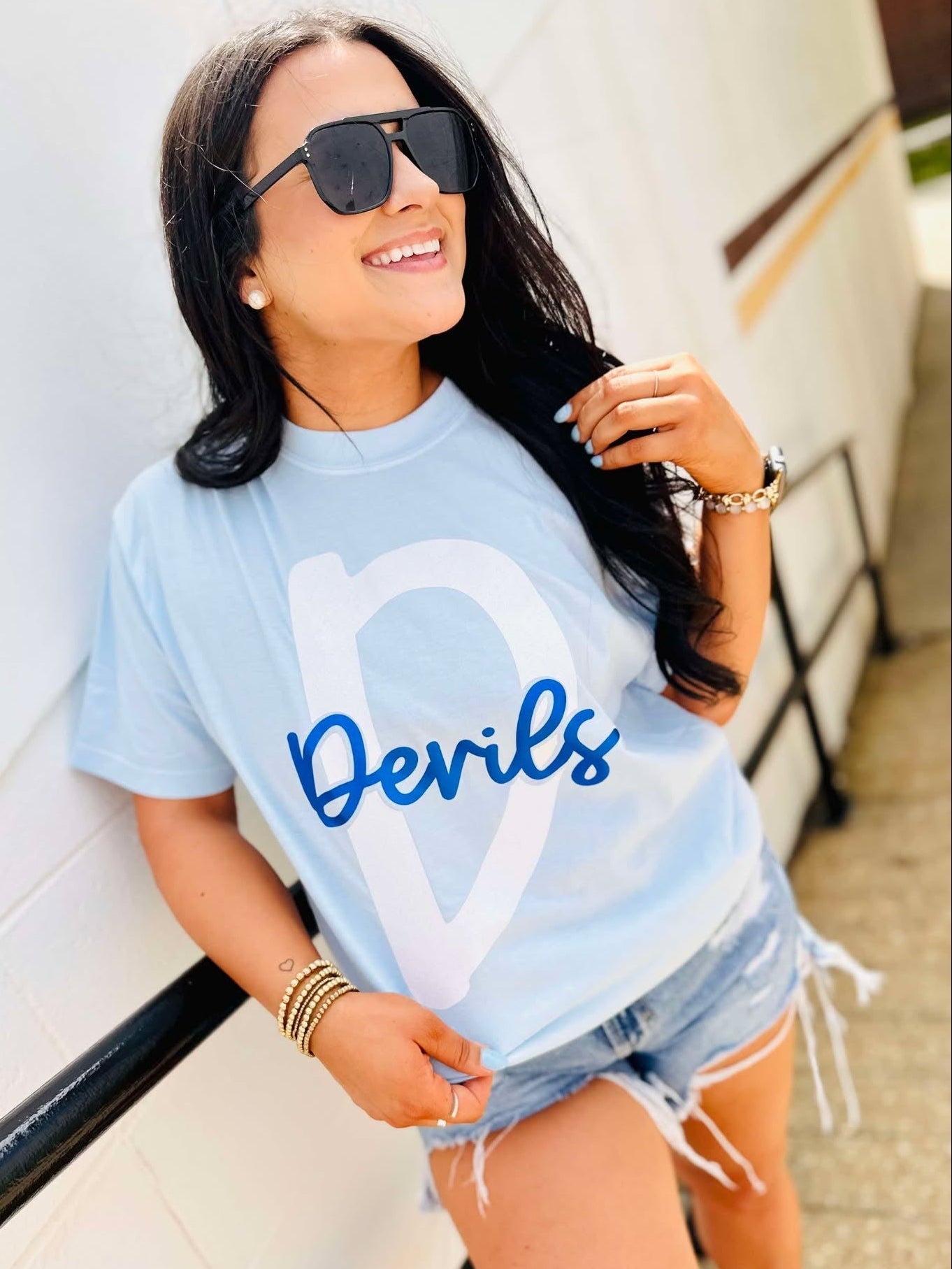 Woman wearing a light blue t-shirt with 'Devils' printed on it, standing against a white wall.