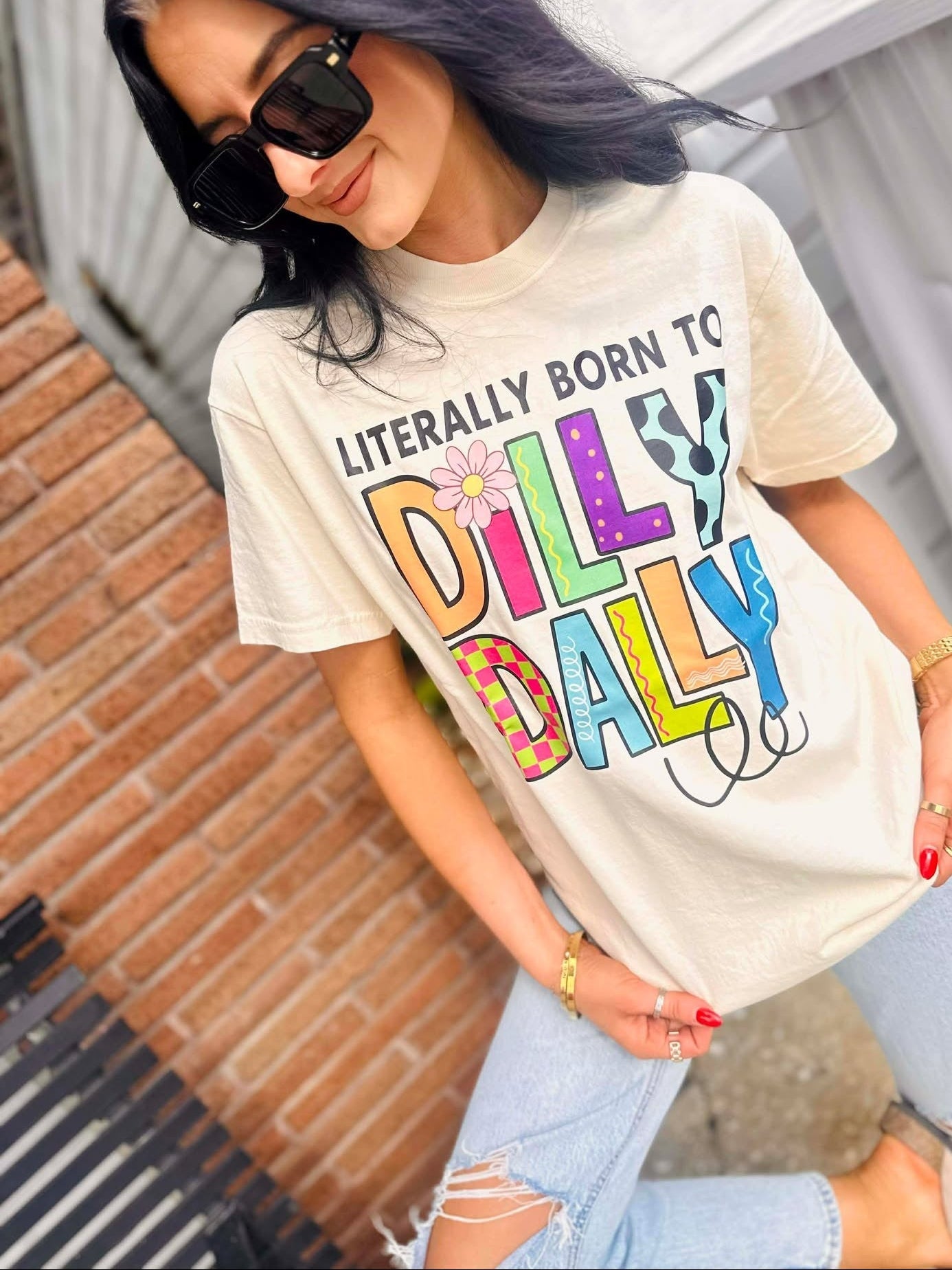 Woman wearing a Born to Dilly Dally Ivory  t-shirt with colorful text outdoors
