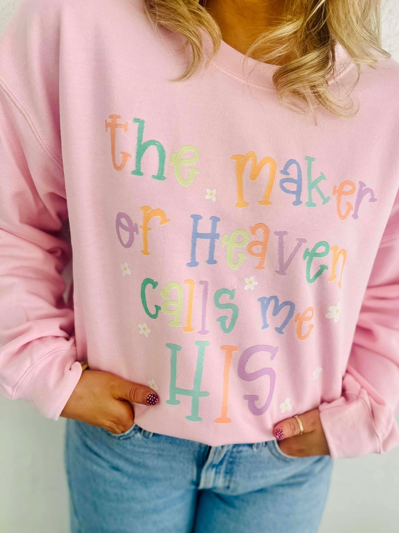 Person wearing a pink sweatshirt with colorful text design, the maker of heaven calls me HIS, standing against a plain background.