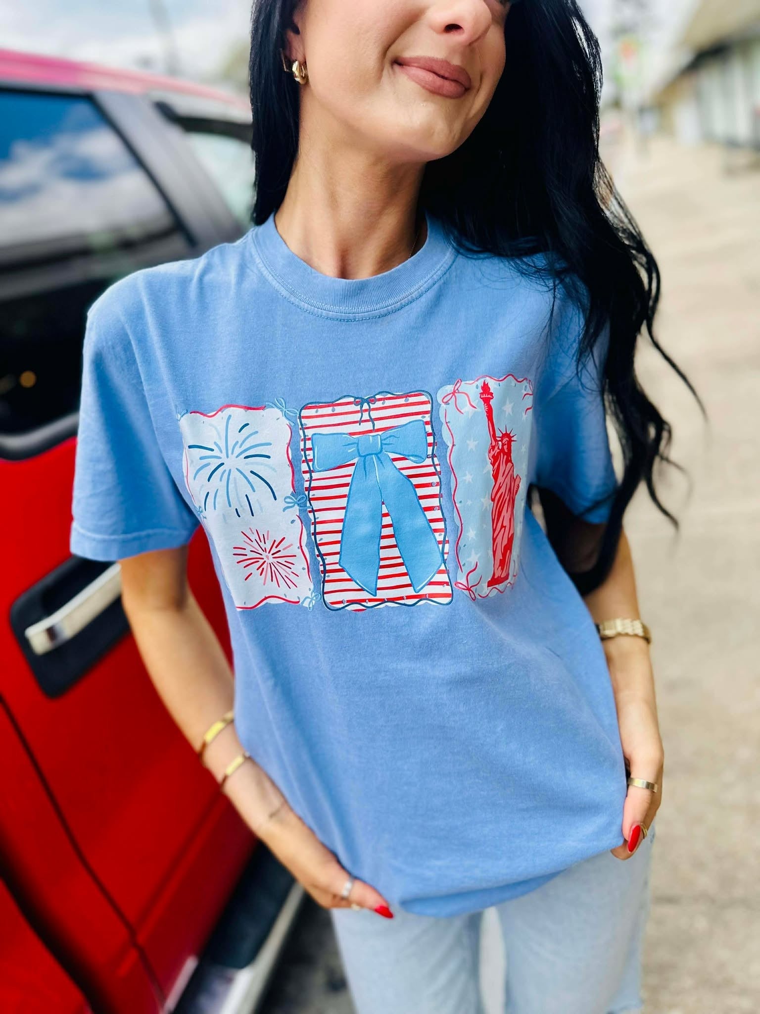 Woman wearing a blue t-shirt with graphic designs, standing next to a red car.