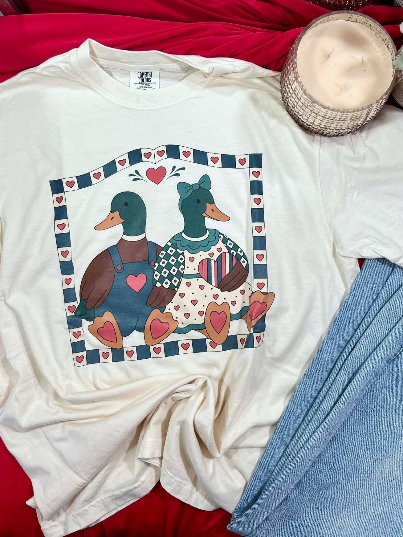 White t-shirt with duck design on a red surface with jeans and candles.
