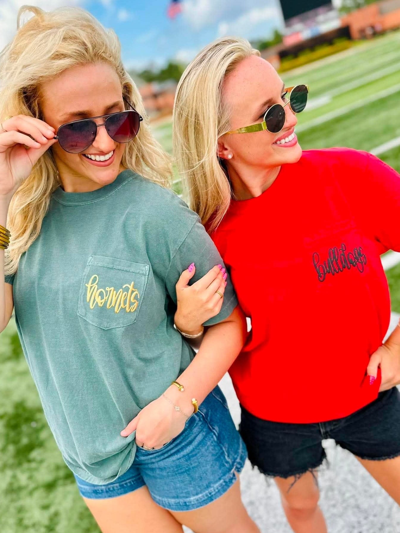 Two women standing on a football field wearing matching graphic t-shirts. The woman on the left wears a muted sage green t-shirt with 'thornte' in gold script on the chest pocket, paired with denim shorts.