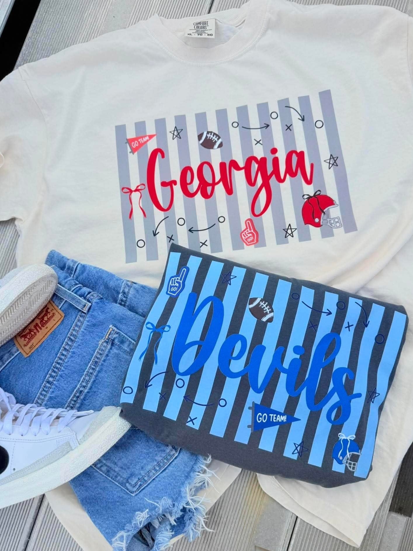 A flat-lay of a white graphic T-shirt with a Georgia Bulldogs sports design, styled with denim shorts, a matching blue striped sweatshirt, and white sneakers on a gray wood plank surface.