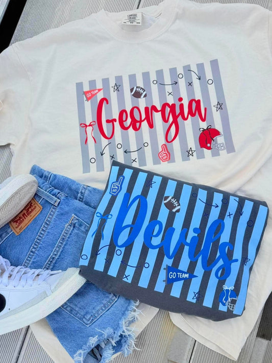A flat-lay of a white graphic T-shirt with a Georgia Bulldogs sports design, styled with denim shorts, a matching blue striped sweatshirt, and white sneakers on a gray wood plank surface.