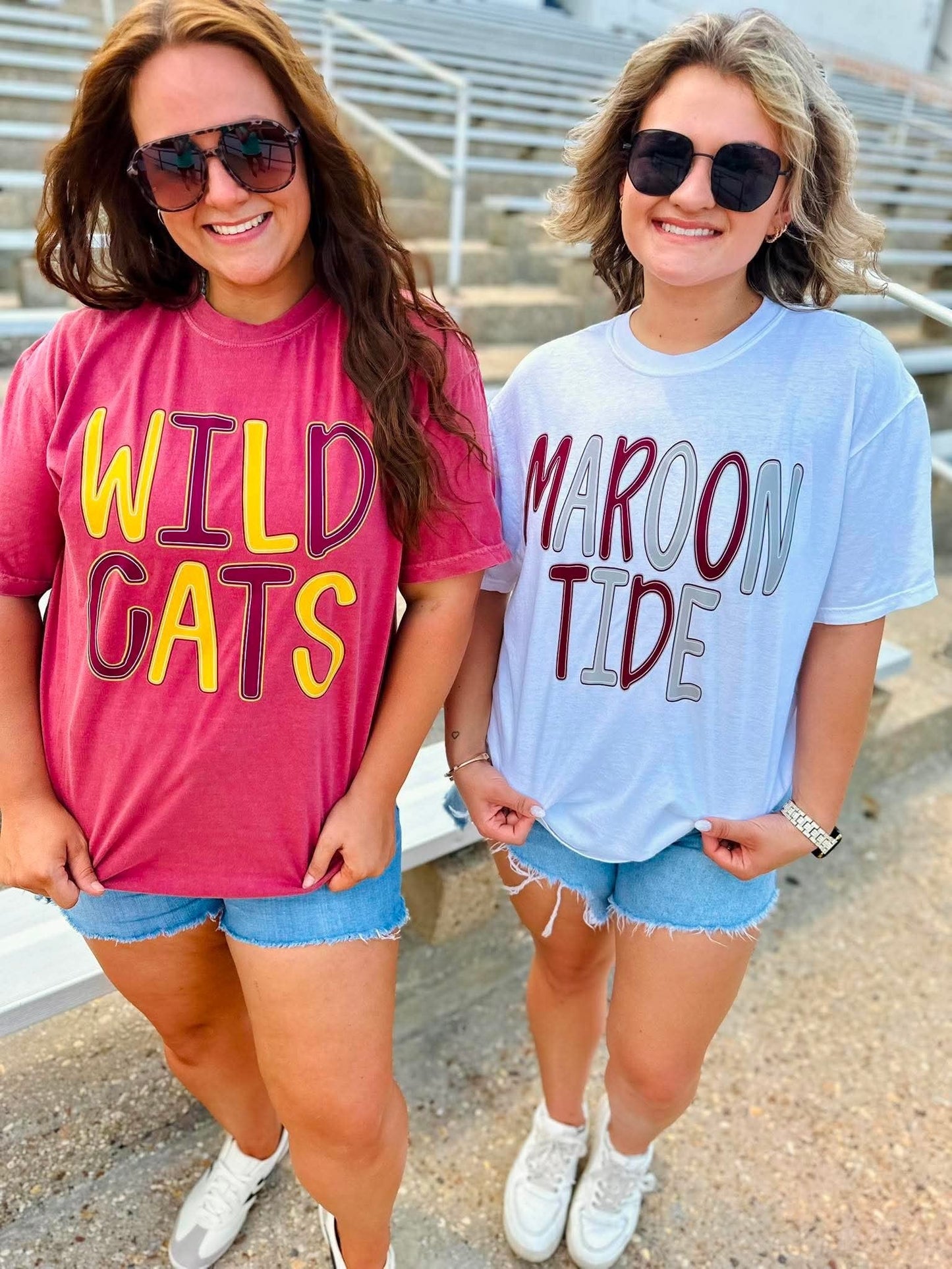 Two women standing outdoors in front of stadium seating, wearing oversized graphic t-shirts. The woman on the left wears a pink t-shirt with 'WILD CATS' in yellow and purple.