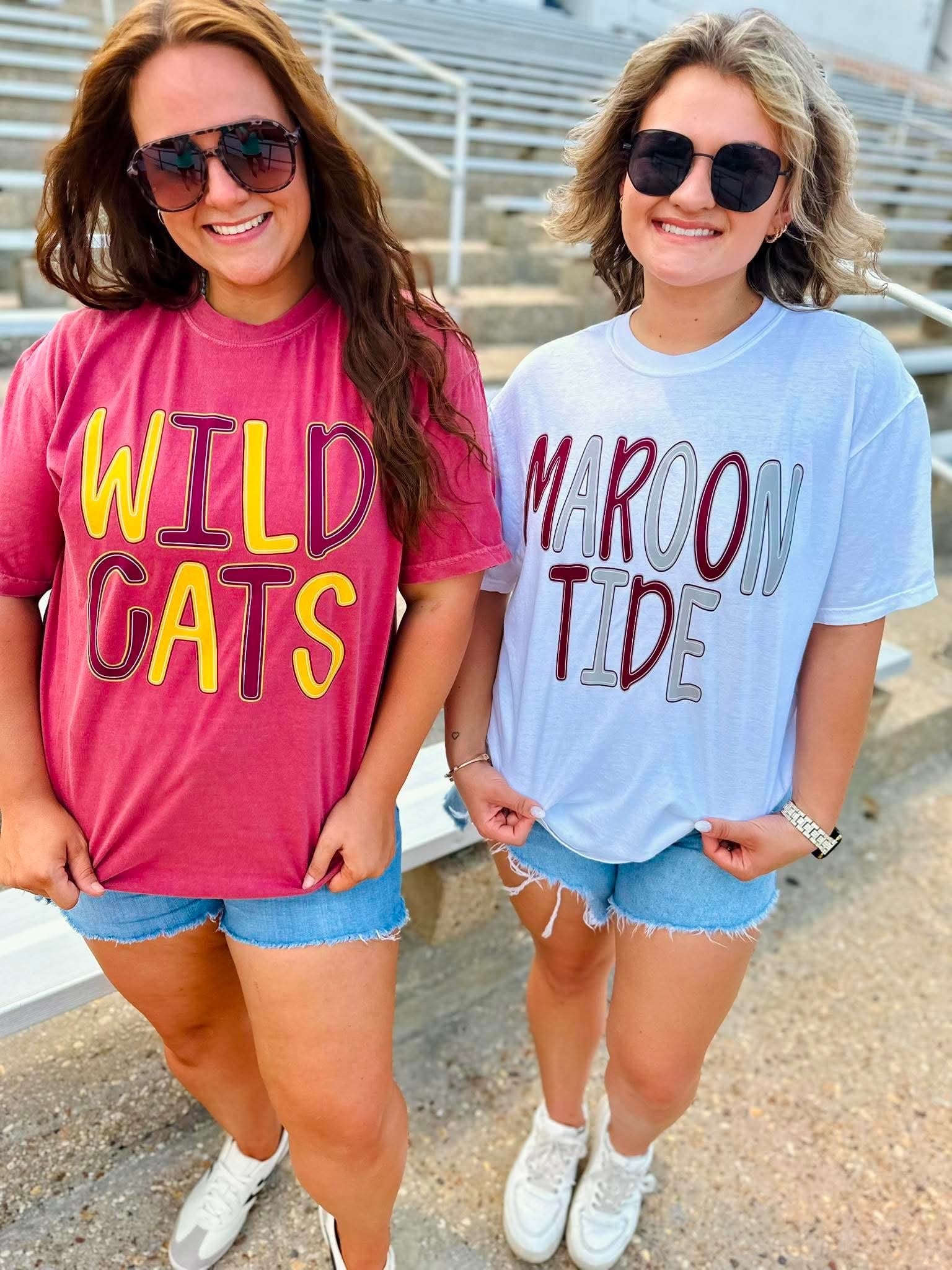 Two women standing outdoors in front of stadium seating, wearing oversized graphic t-shirts. The woman on the left wears a pink t-shirt with 'WILD CATS' in yellow and purple.