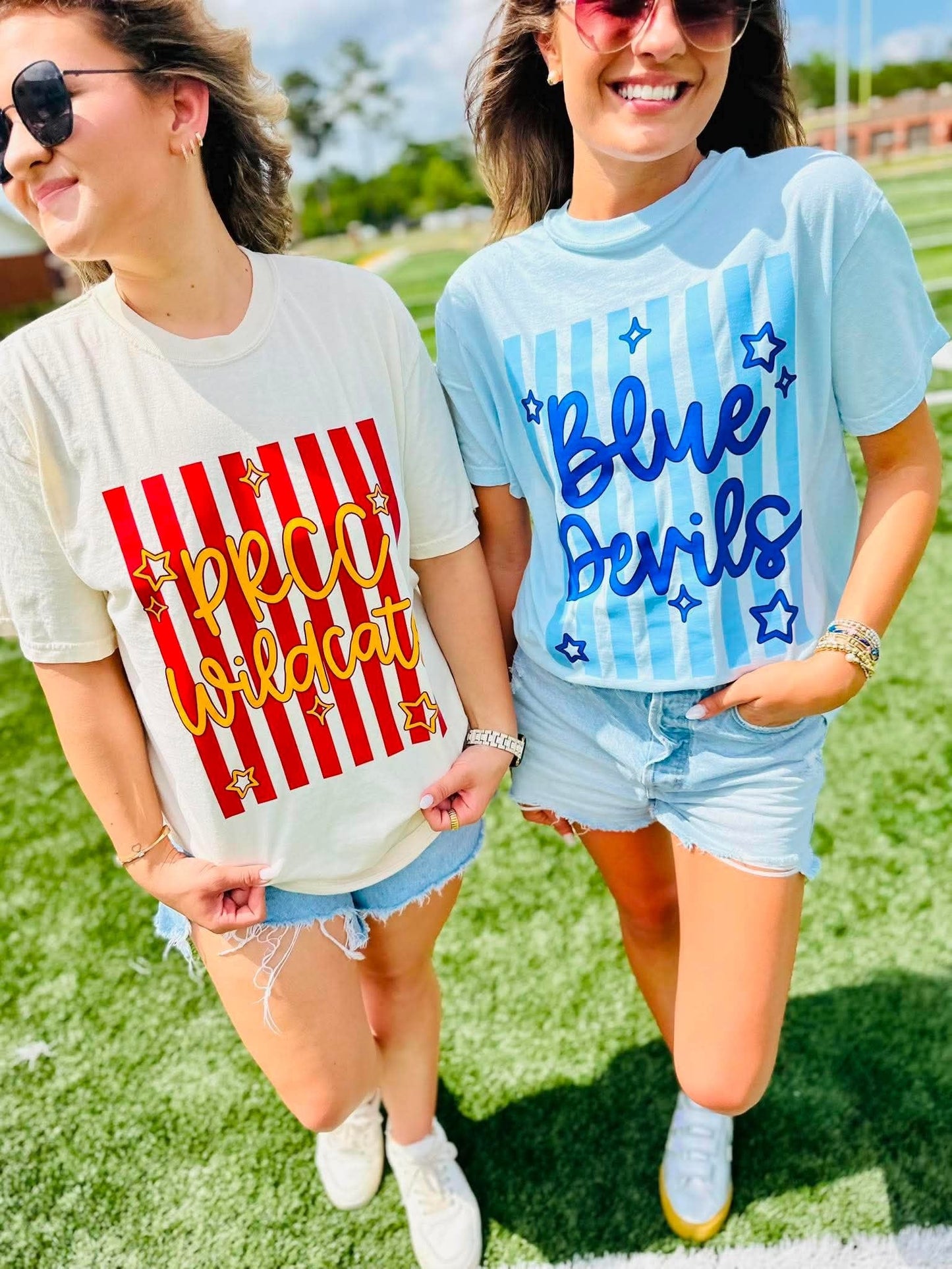 Two women walking outdoors on a grassy field wearing graphic t-shirts. The woman on the left wears a cream t-shirt with red and yellow 'PRCC Wildcat' graphics, and the woman on the right wears a light blue t-shirt with 'Blue Devils' and blue stars.