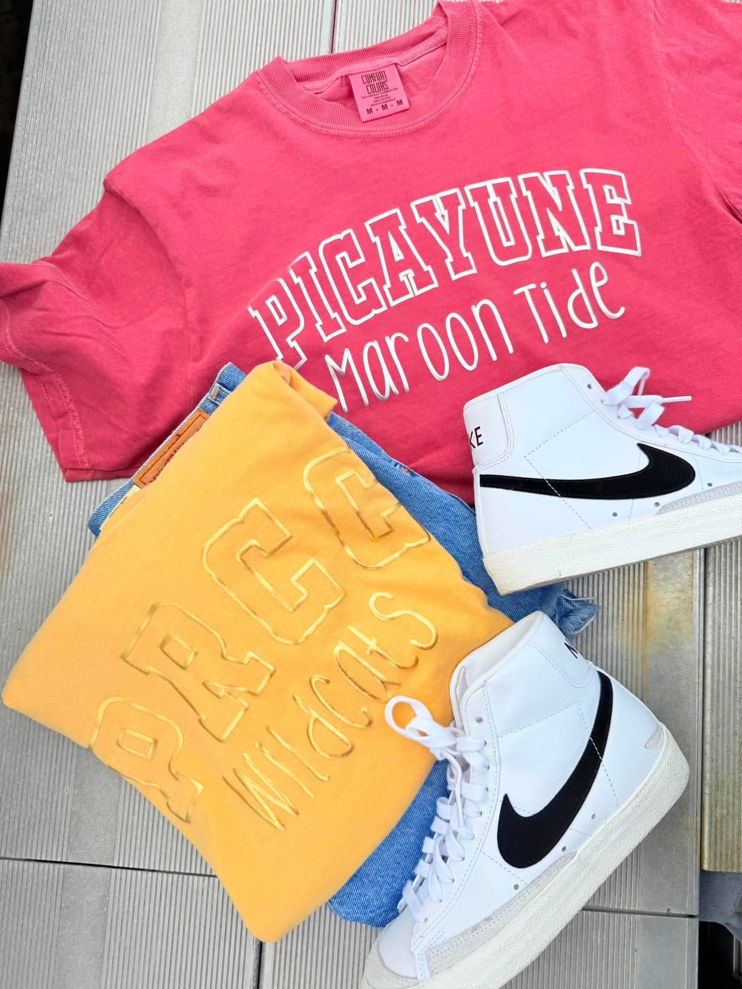 Pink t-shirt with 'PICAYUNE Maroon Tide', yellow towel, blue jeans, and white sneakers on a wooden floor.