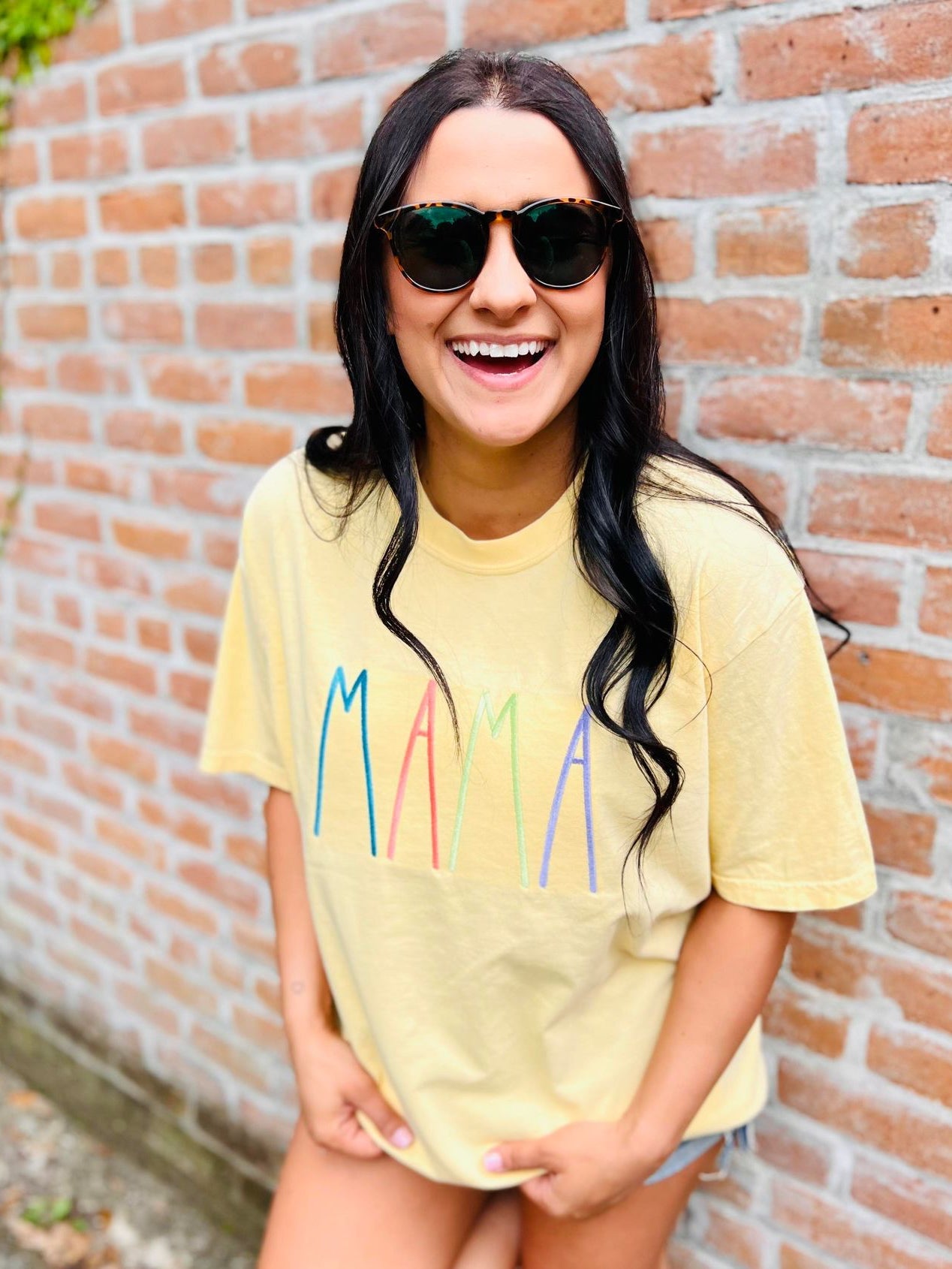 A person wearing a yellow graphic tee with the text 'MAMA' embroidered on the front, standing with one hand against a brick wall.