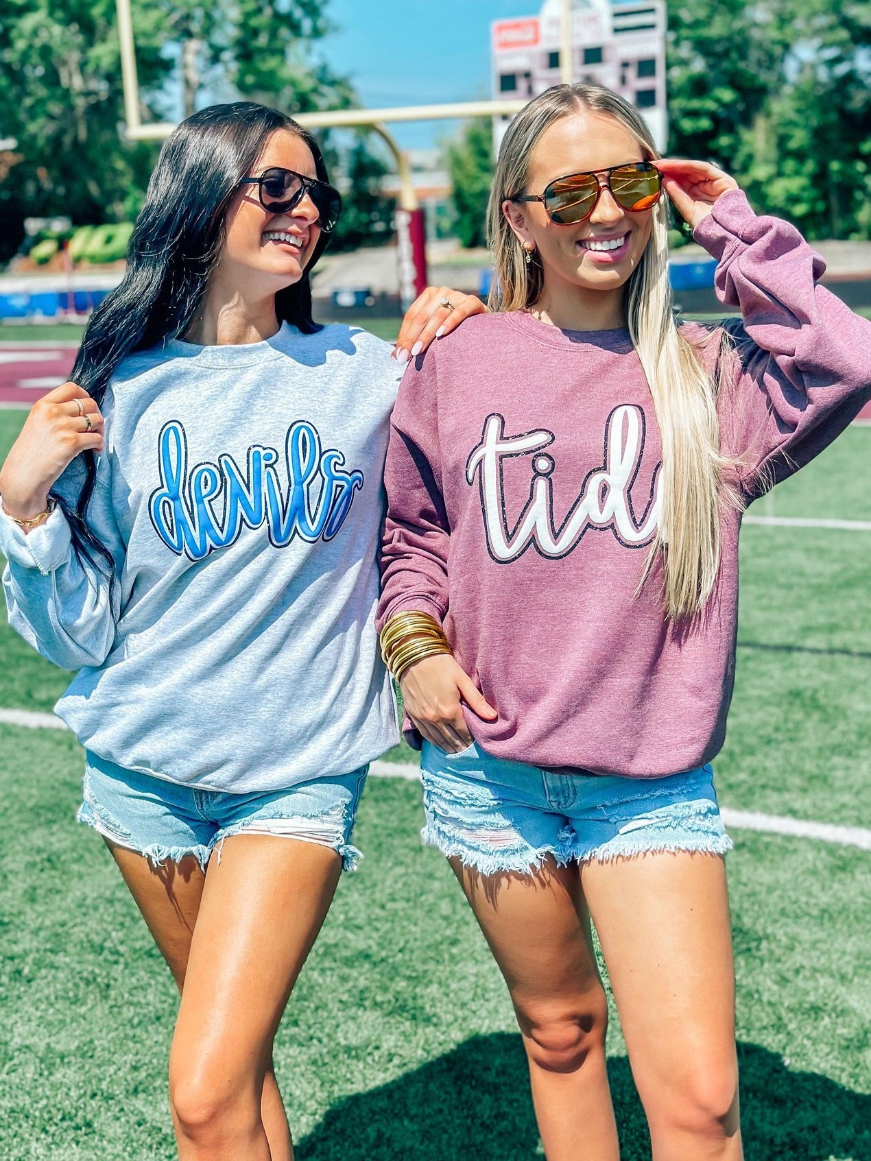 Two women standing on a field wearing sunglasses and custom mascot sweatshirts in blue and gray, with one woman's shirt displaying the word 'DEVILS' and the other's shirt displaying the word 'TIDE'.