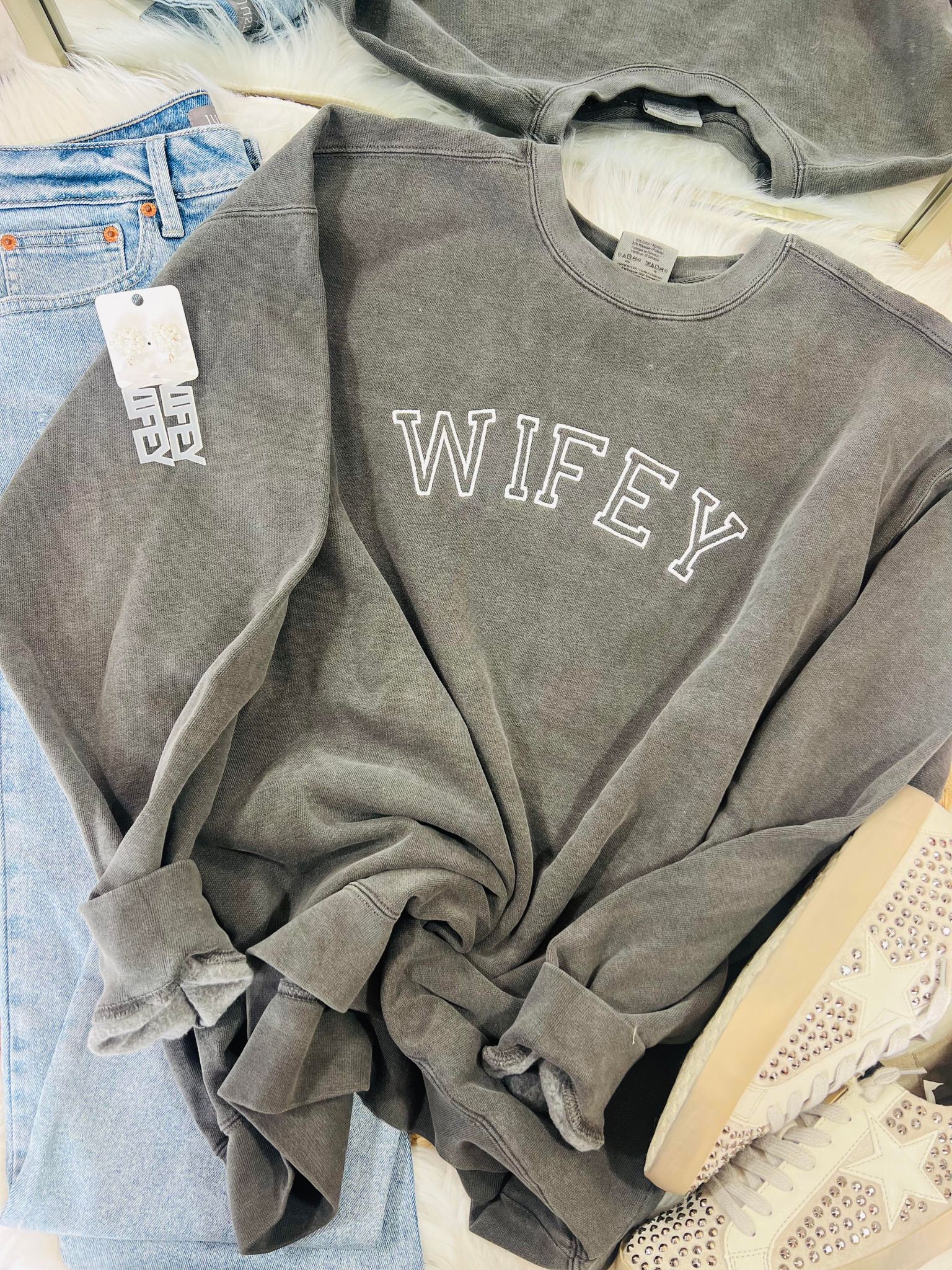 A gray sweatshirt with the word 'WIFEY' embroidered in white on the front, displayed on a rack with other clothing items.