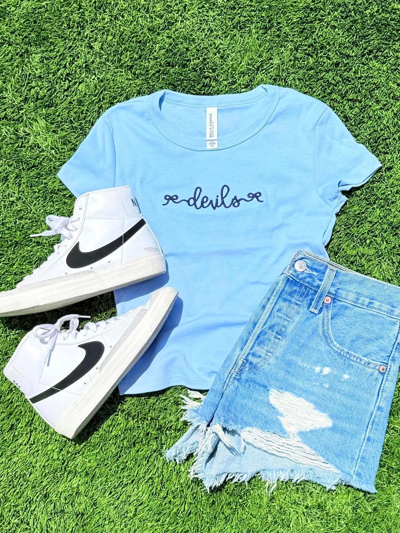 Light blue t-shirt with 'devils' text, white high-top sneakers, and denim shorts on grass