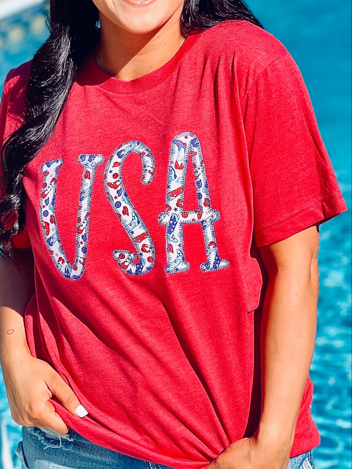 A person wearing a red t-shirt with the text 'USA' and an embroidered design resembling a flag on it, standing by a pool.