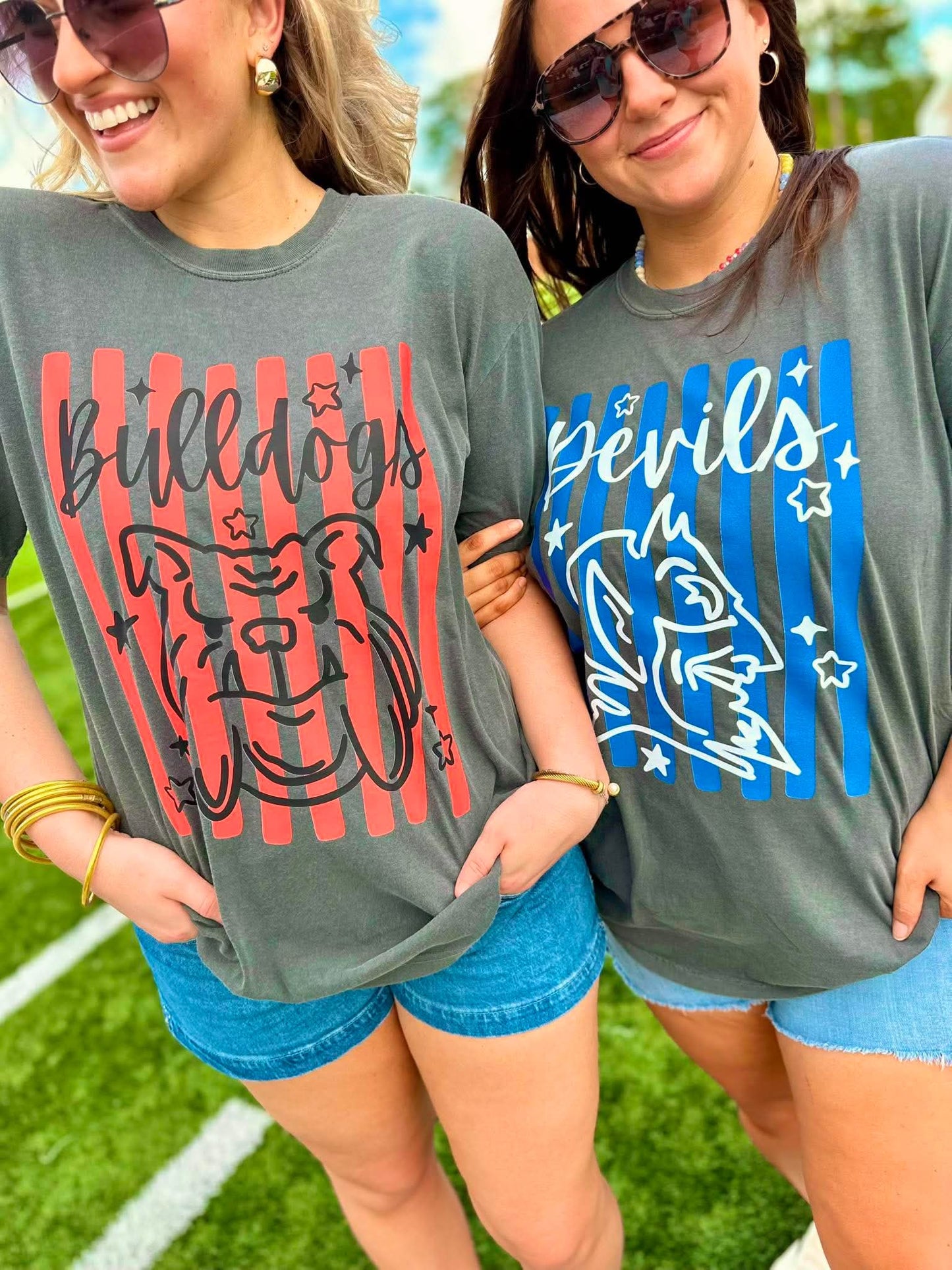 Two women wearing gray t-shirts with 'Bulldogs' and 'Devils' graphics on a grassy field.
