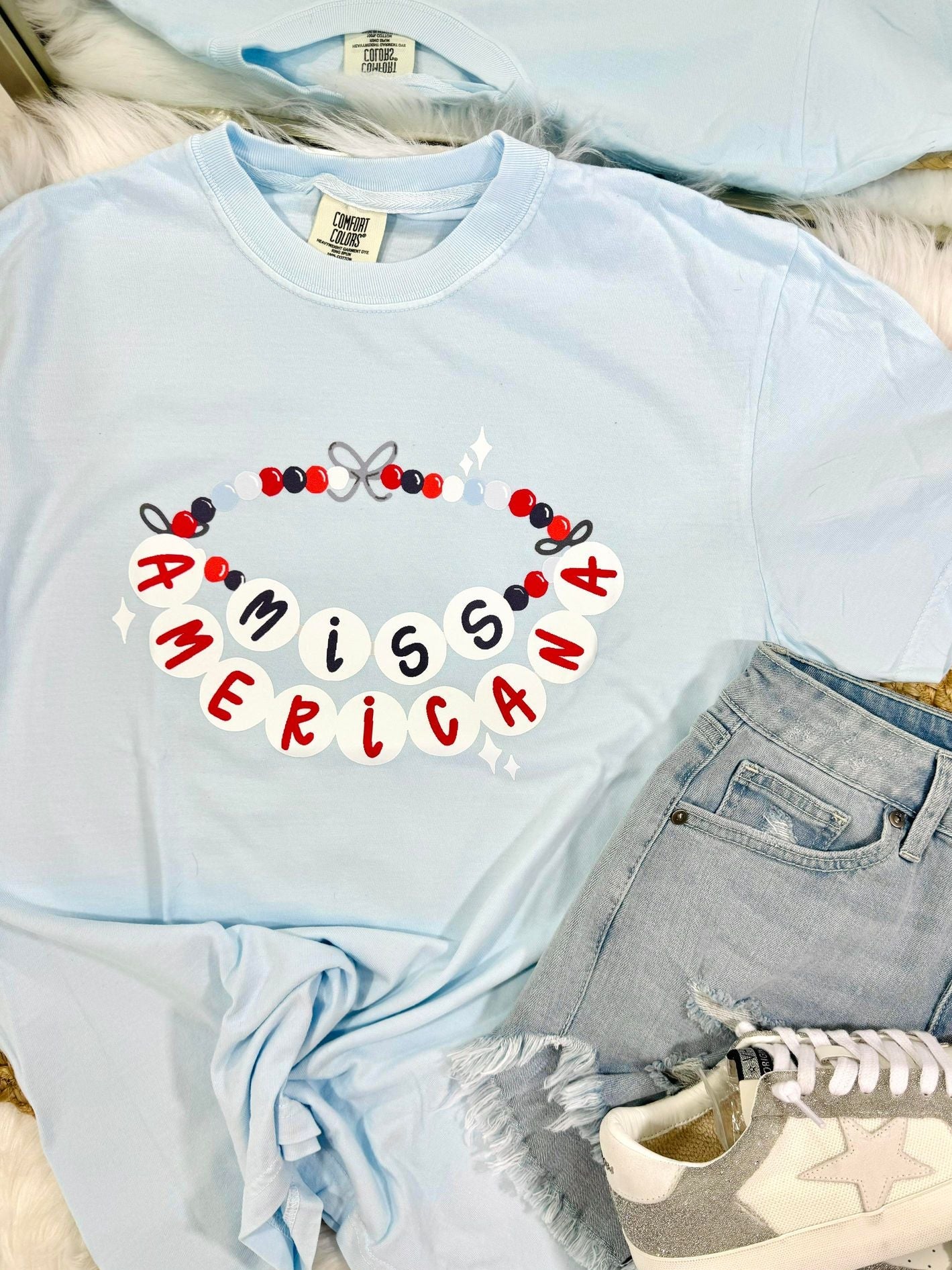 A light blue t-shirt with the text 'MISS AMERICA' and decorative friendship bracelet elements printed across the front, displayed flat with denim shorts and white sneakers.