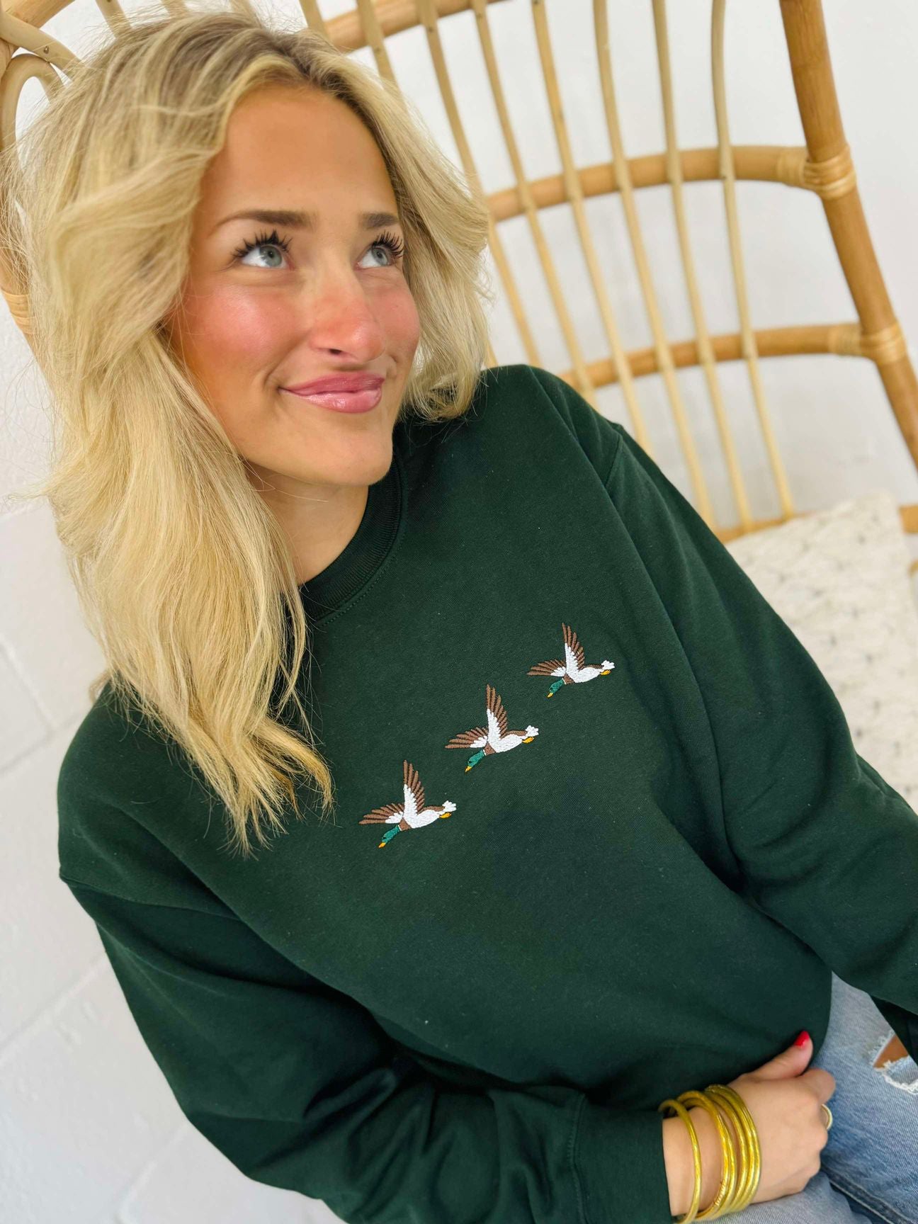 A woman sitting on a wooden chair wearing a green embroidered sweatshirt with a trio of mallard ducks on the front.