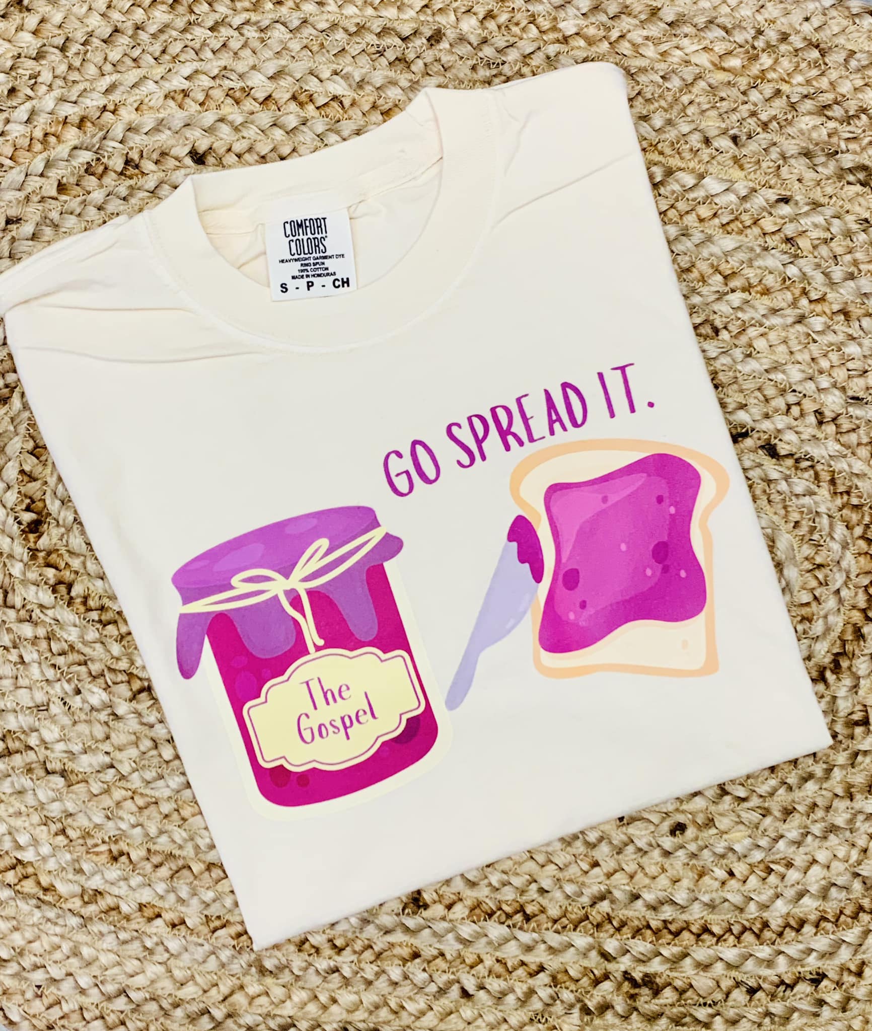 A youth-sized white t-shirt with a graphic print of a jar and a slice of bread, alongside the phrase 'GO SPREAD IT.'