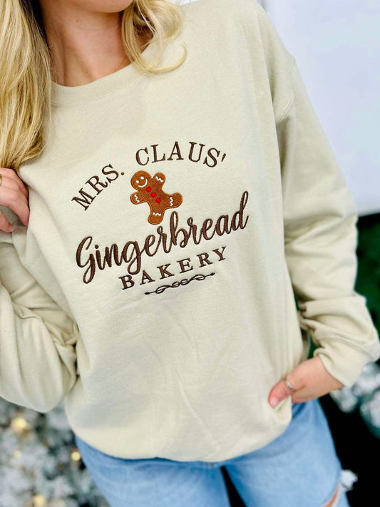 A woman wearing a beige embroidered sweatshirt with 'MRS. CLAUS' GINGERBREAD BAKERY' text and a cute gingerbread man graphic.