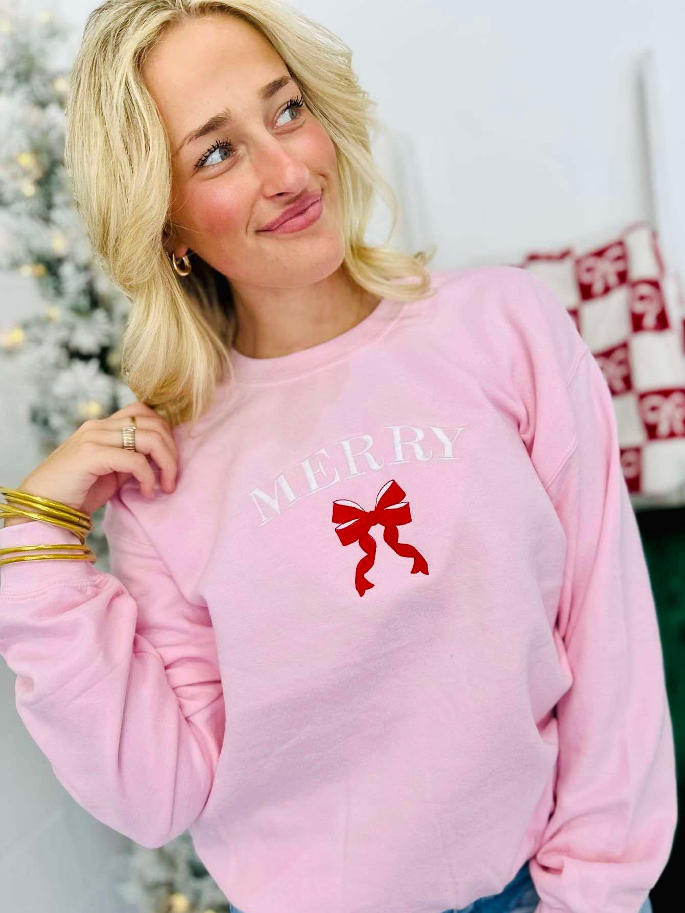 A woman wearing a light pink sweatshirt with 'Merry' embroidered on the front and a red bow detail.