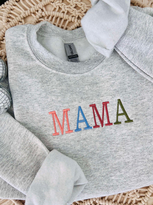 A gray sweatshirt with 'Mama' embroidered in red and green colors on the front, displayed in a woven basket with decorative balls and a tag.